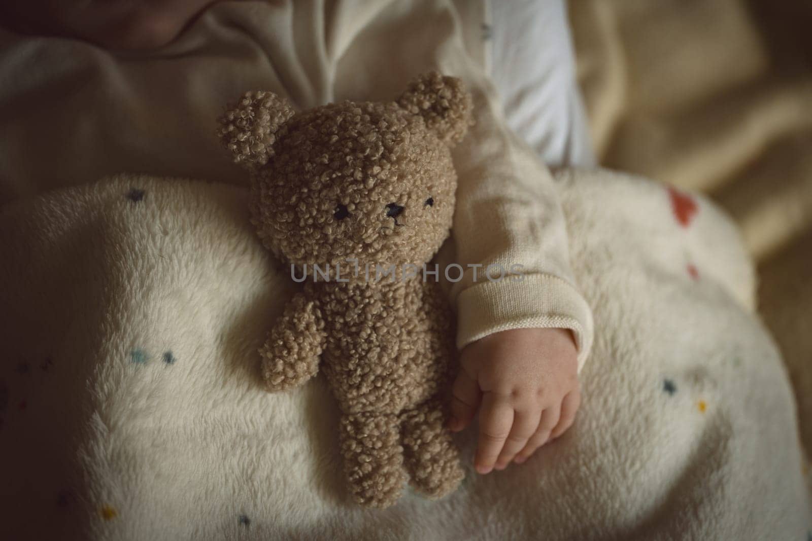 Fluffy toy teddy bear and baby little hand lying in bed in warm, cozy sunlight. Childhood and parenting concept. High quality photo