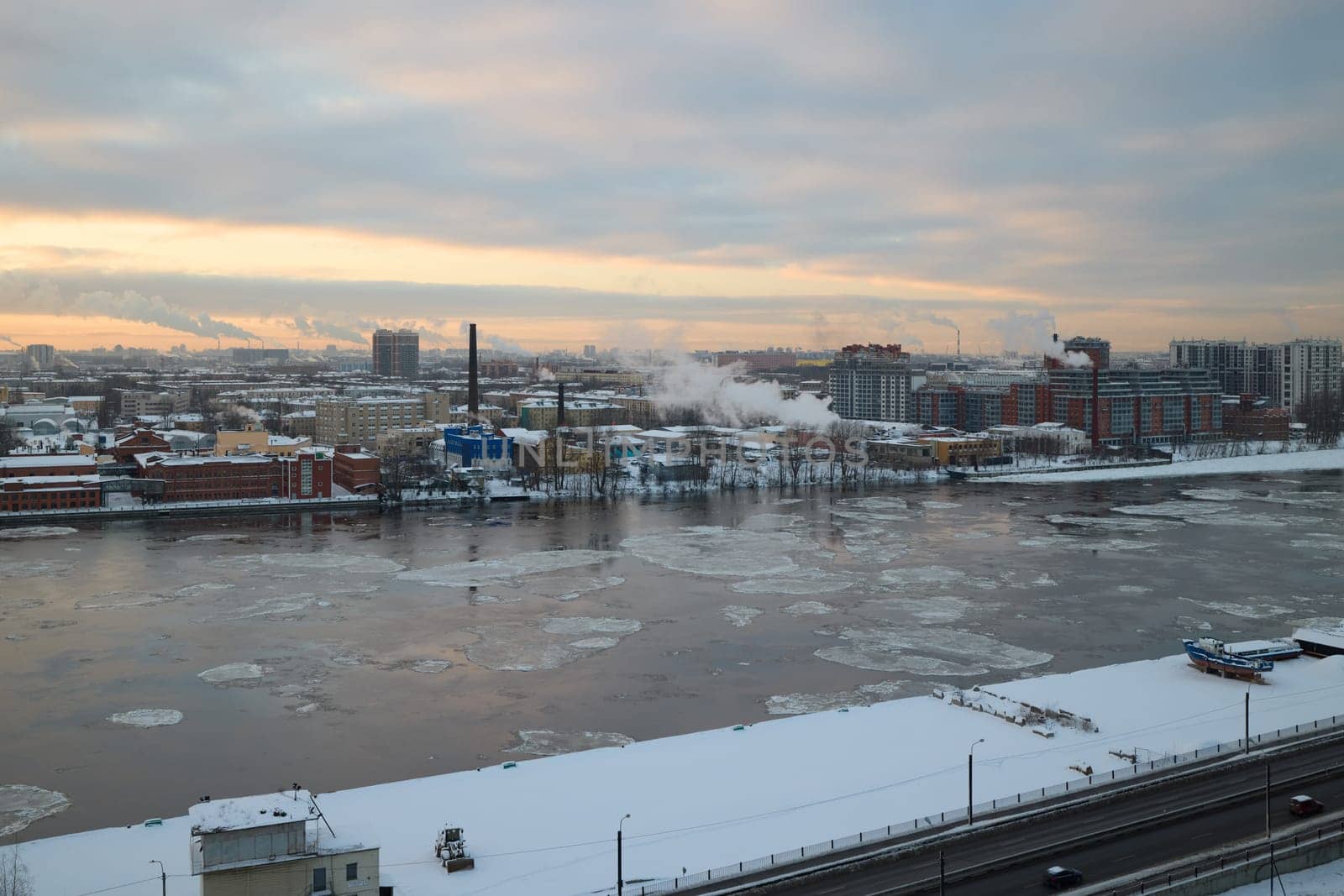 A serene winter landscape showcasing a river with ice, smoke, and city buildings against a cloudy sky.