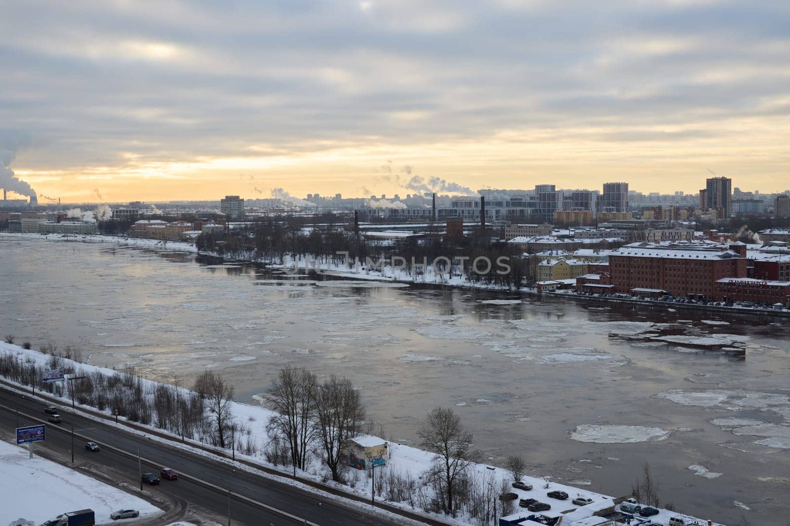 A serene winter view of a tranquil river, surrounded by snow, buildings, and a cloudy sky at sunset time