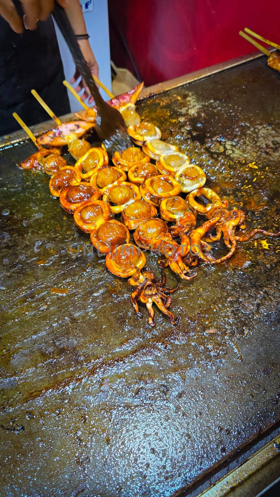 cook grills skewered squid over an open flame using a blowtorch, capturing the lively atmosphere of a bustling street food Thai market with bright flames and freshly prepared seafood.