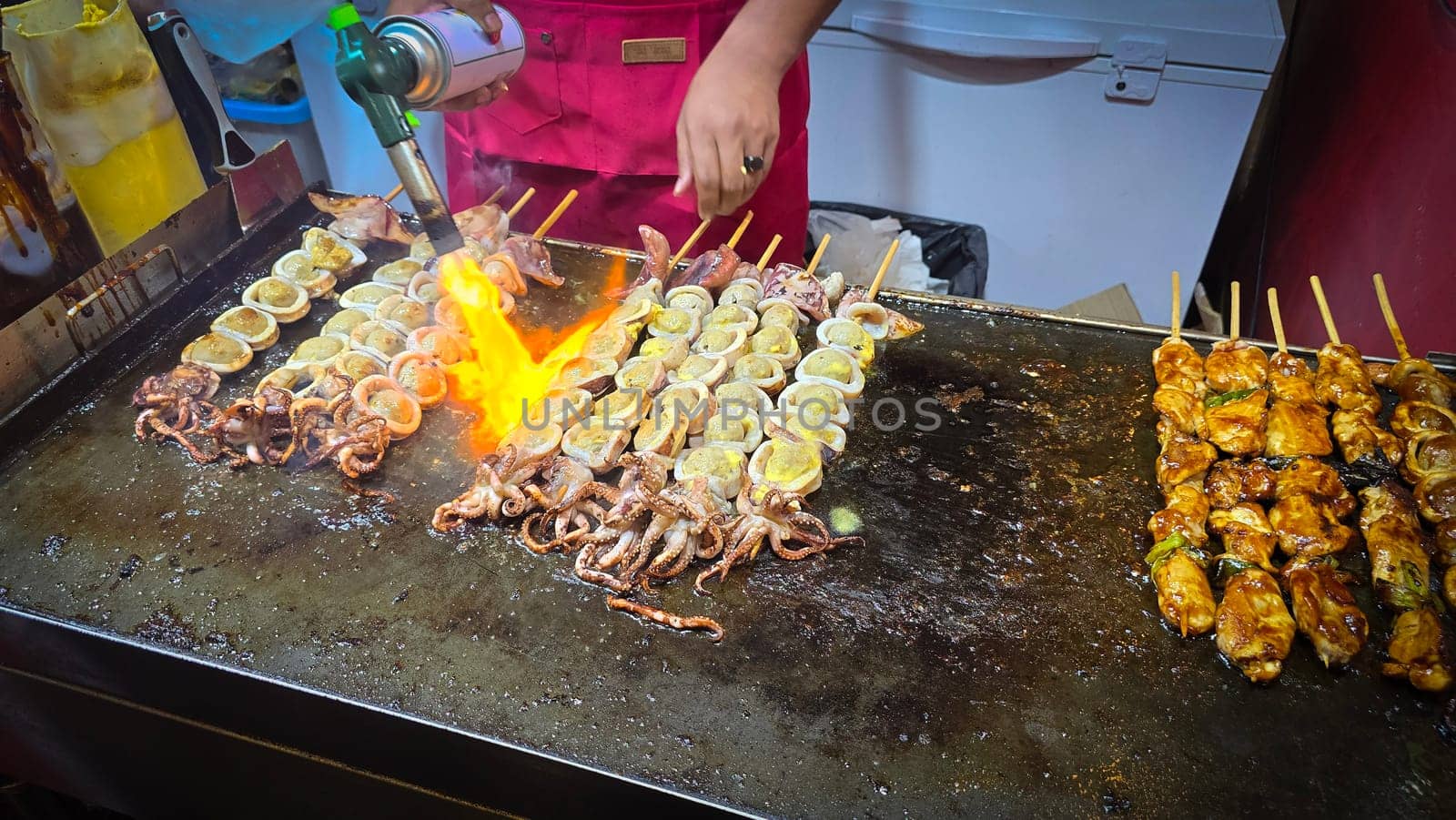 cook grills skewered squid over an open flame using a blowtorch, capturing the lively atmosphere of a bustling street food Thai market with bright flames and freshly prepared seafood.