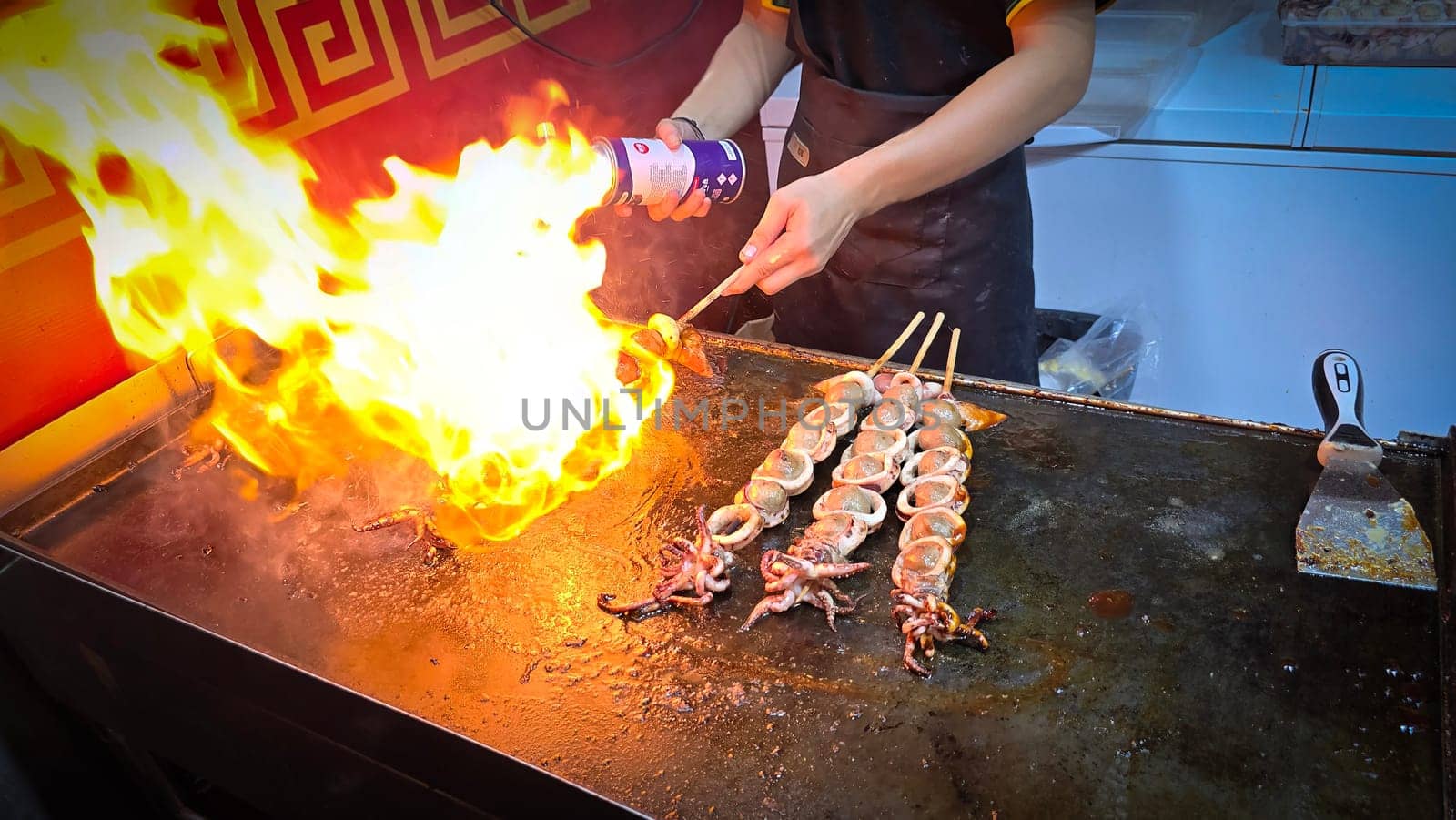 cook grills skewered squid over an open flame using a blowtorch, capturing the lively atmosphere of a bustling street food Thai market with bright flames and freshly prepared seafood.