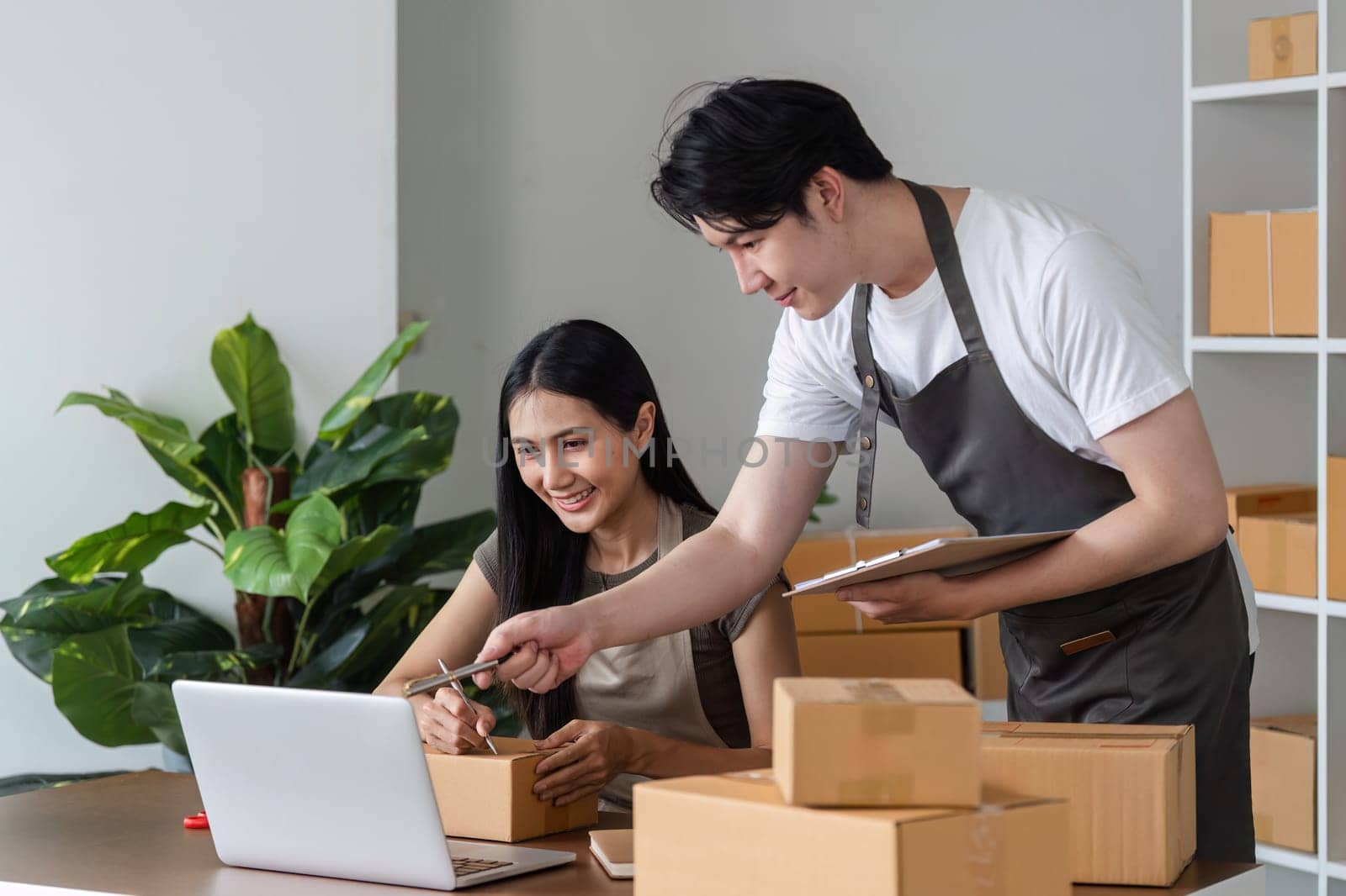 A couple collaborates on their online business from home, surrounded by shipping boxes and using a laptop in a modern, organized workspace.