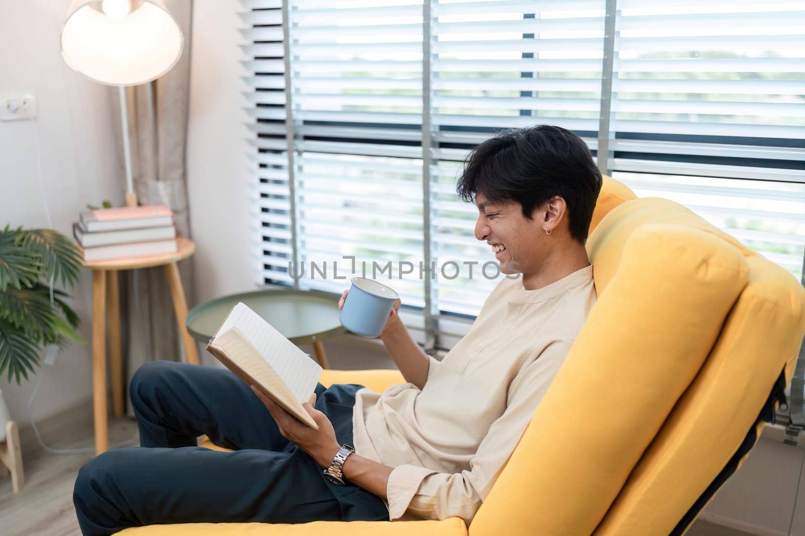 A young man sits comfortably in a modern living room, reading a book and holding a hot beverage, creating a cozy and relaxed atmosphere.
