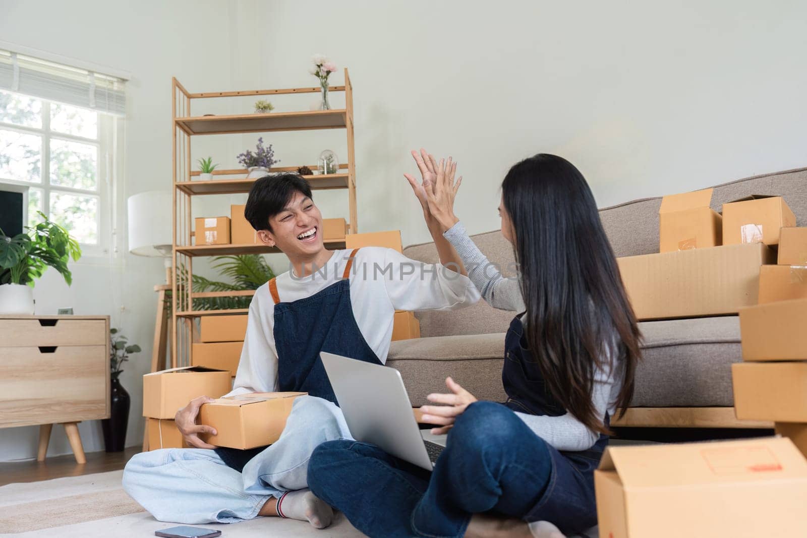 A joyful couple celebrates their successful online sales with a high five, sitting in a modern living room surrounded by shipping boxes and a laptop.