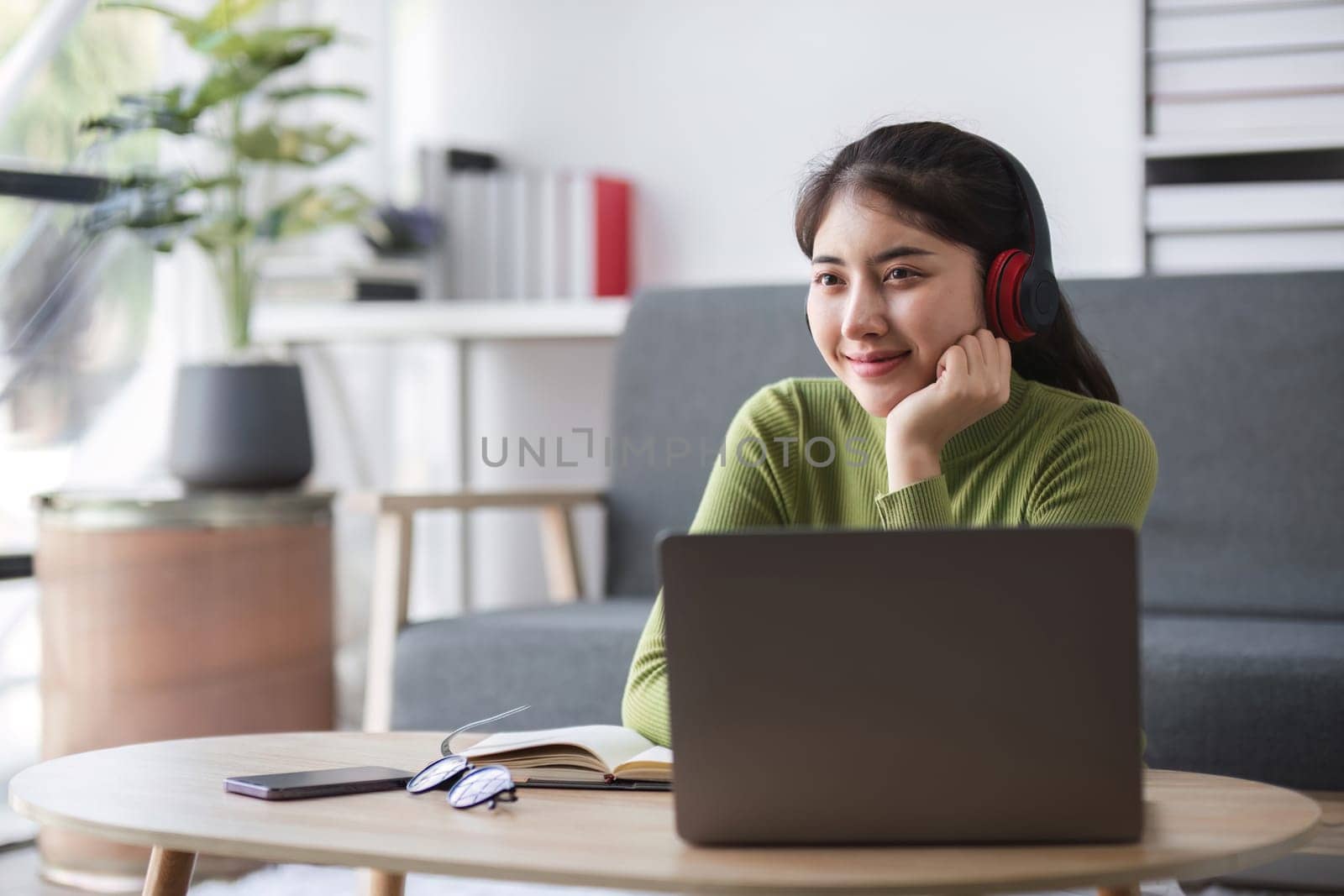 A young woman studies online from home, using a laptop and headphones, seated in a modern living room with a cozy atmosphere.