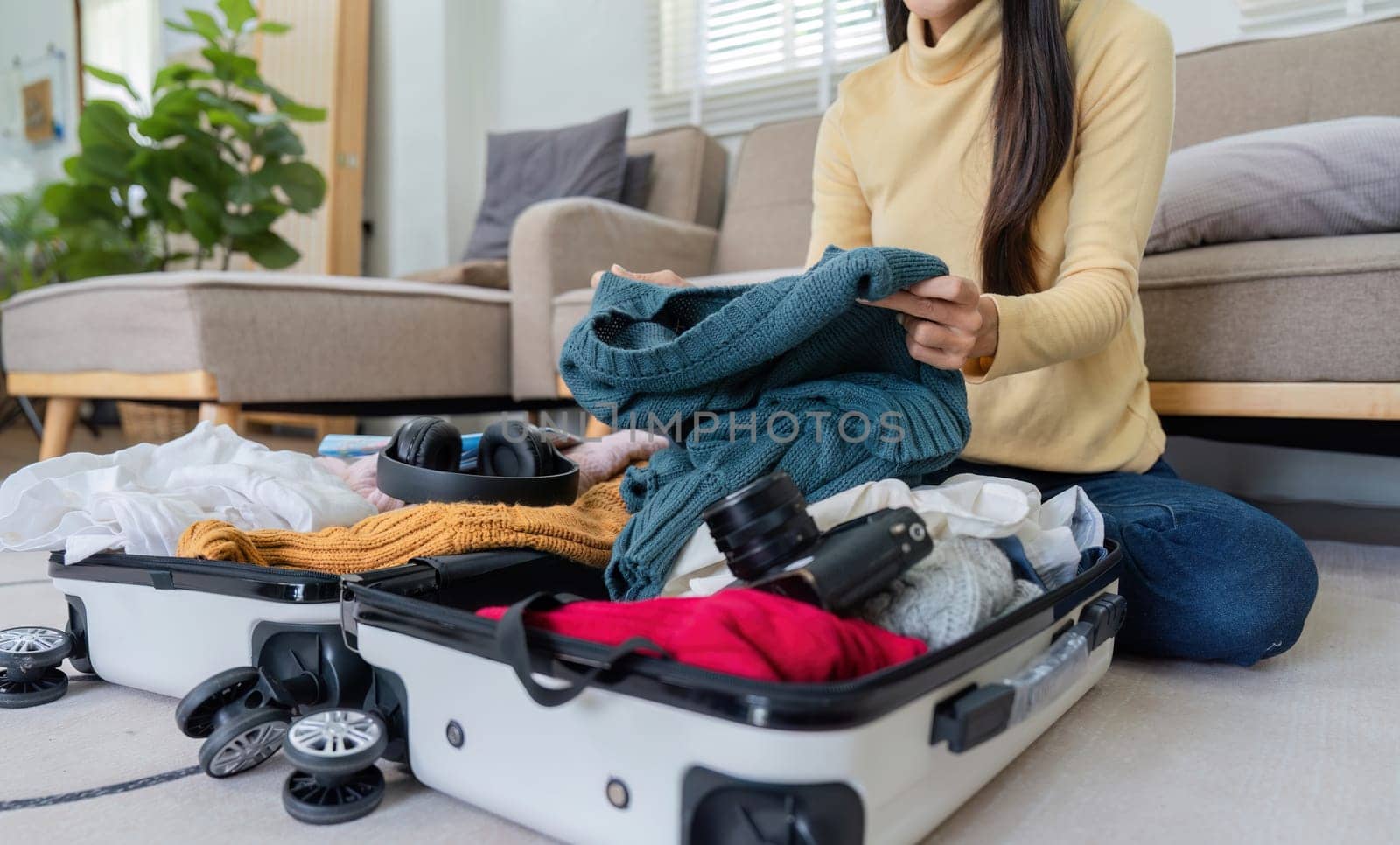A young woman packs her suitcase in a modern living room, organizing clothes and accessories for travel or vacation.