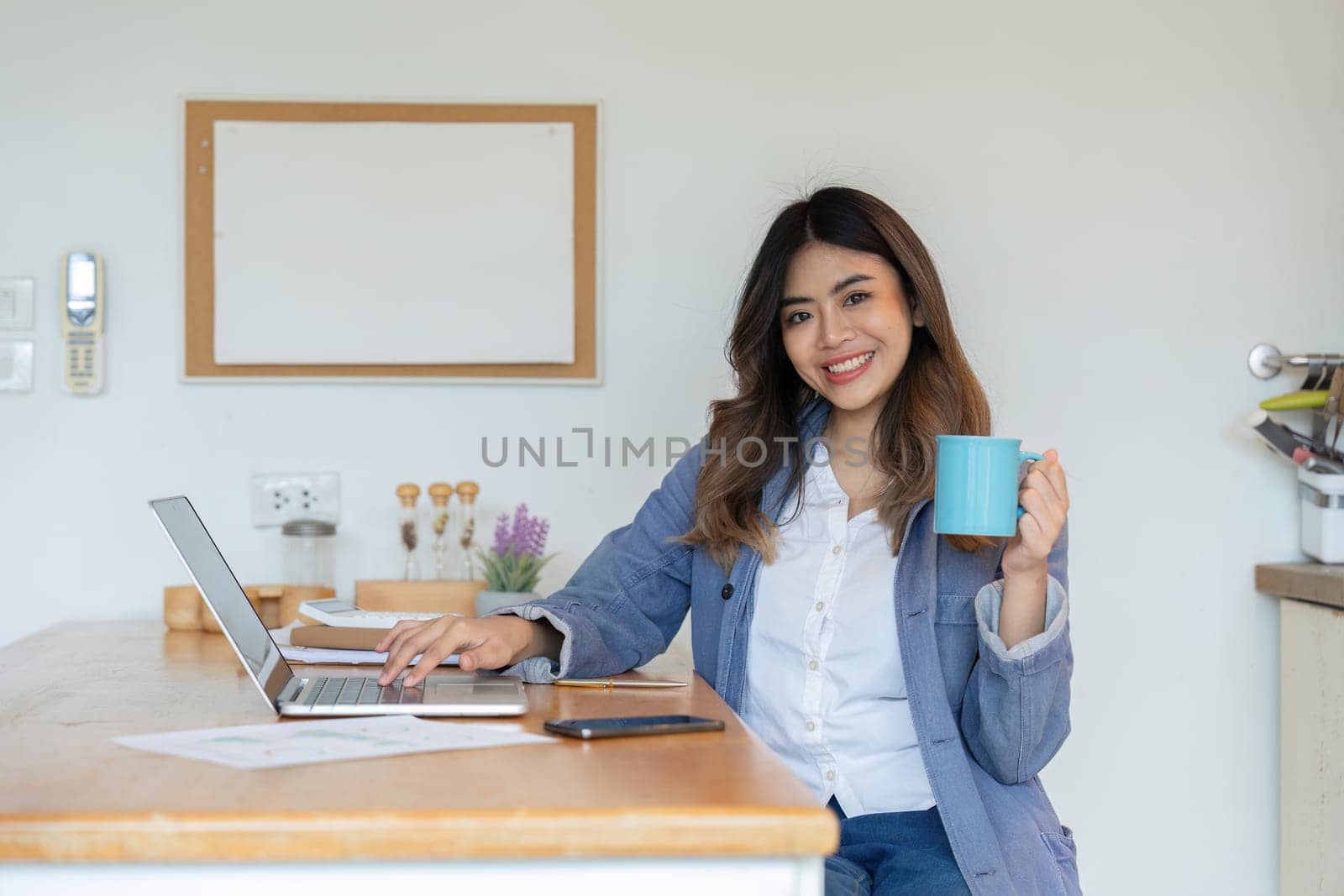 A young woman working from home in a dining room, using a laptop and holding a coffee mug, smiling and appearing productive.