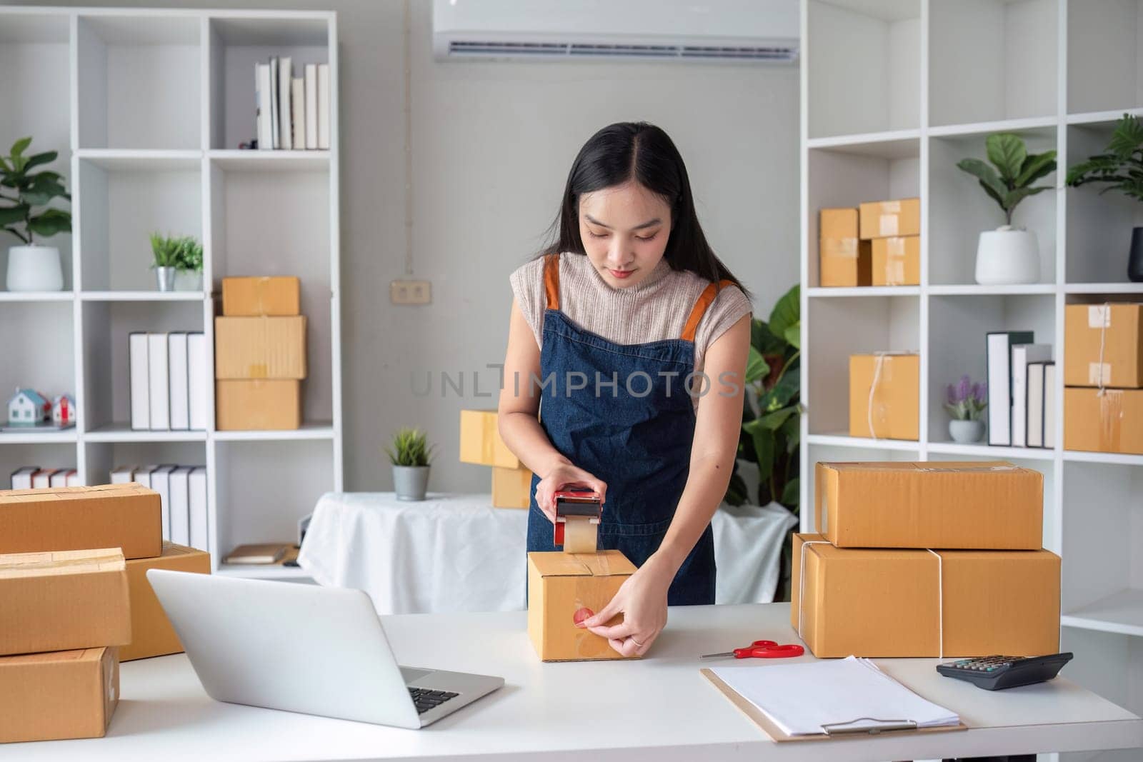 A young female business owner packs products for her online store in a modern home office, surrounded by shipping boxes and a laptop.