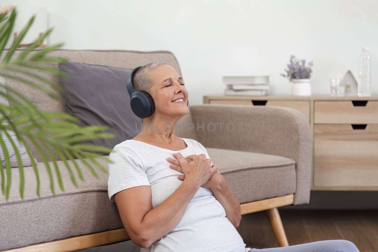 Healthy elderly woman with headphones, eyes closed, enjoying relaxing music in a cozy living room, symbolizing peace and wellness.