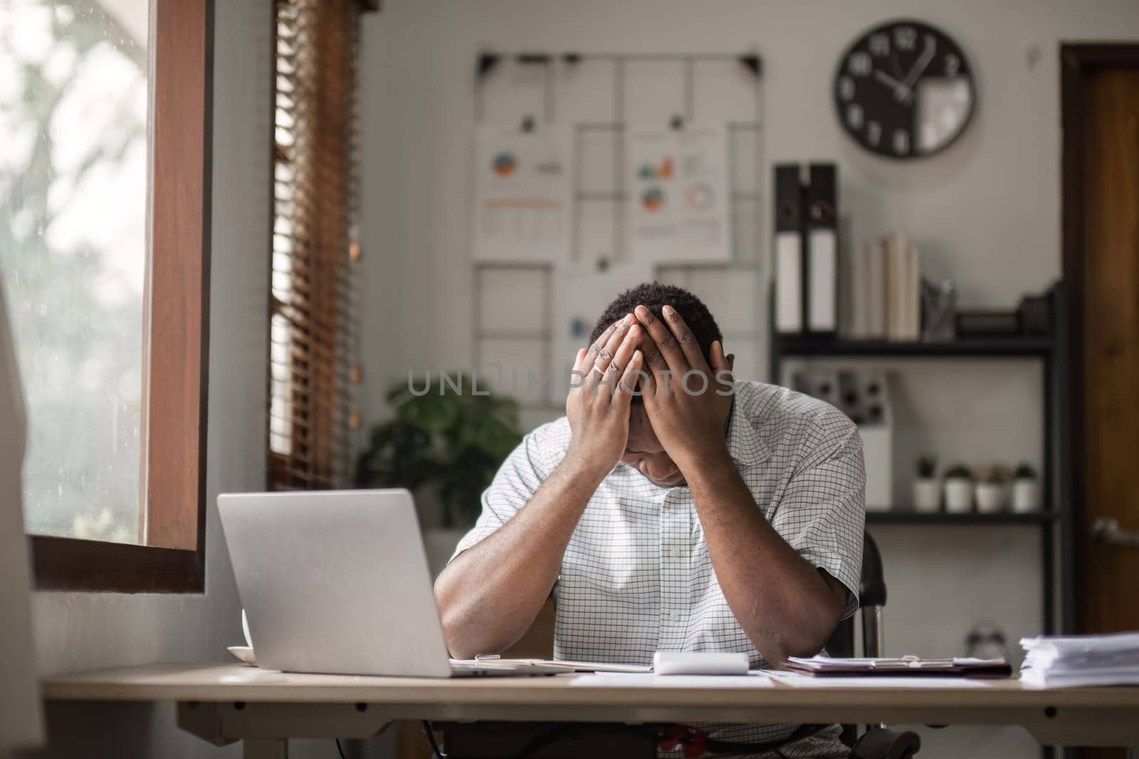 A male office worker experiencing stress and pressure at his desk, surrounded by work materials in a modern office setting.