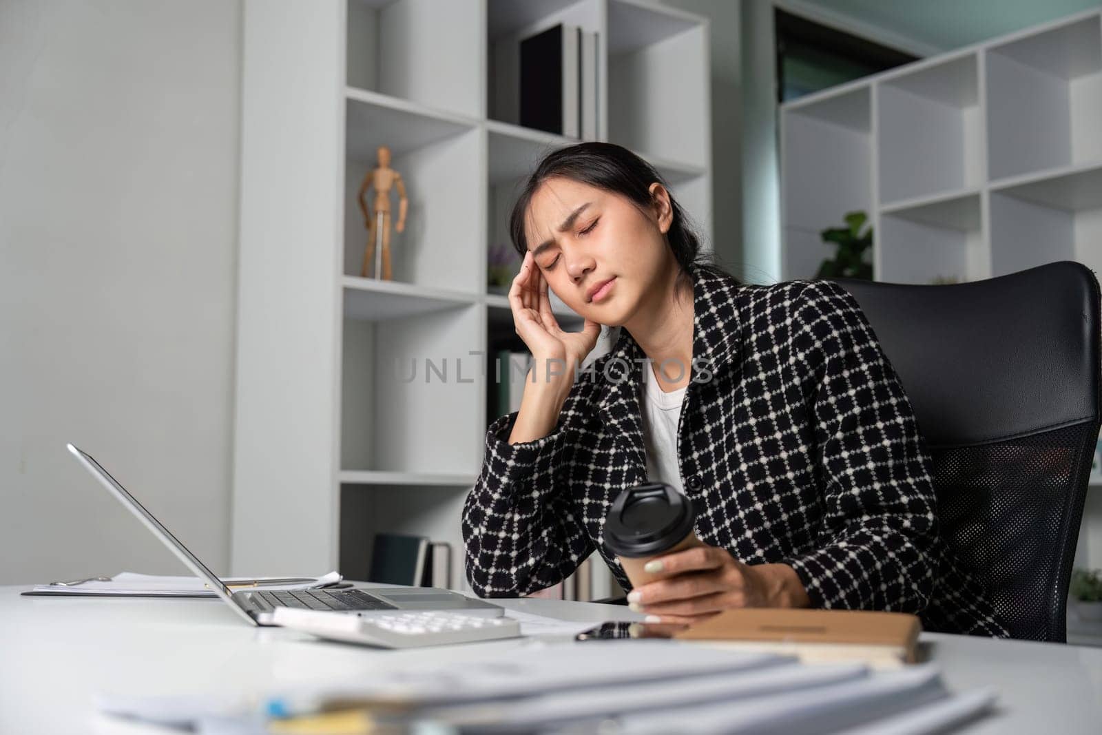 A young businesswoman feeling stressed and overwhelmed from working too much, sitting at her office desk with a laptop and coffee in a modern workspace.