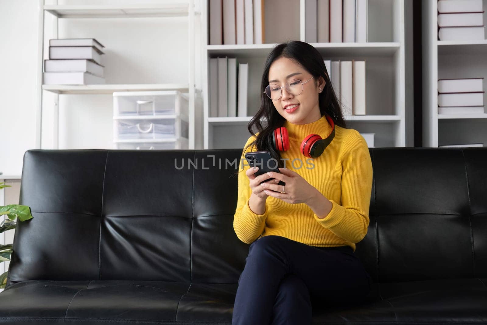 A young woman in a yellow sweater and glasses relaxes happily in a modern living room, using her smartphone and wearing headphones.