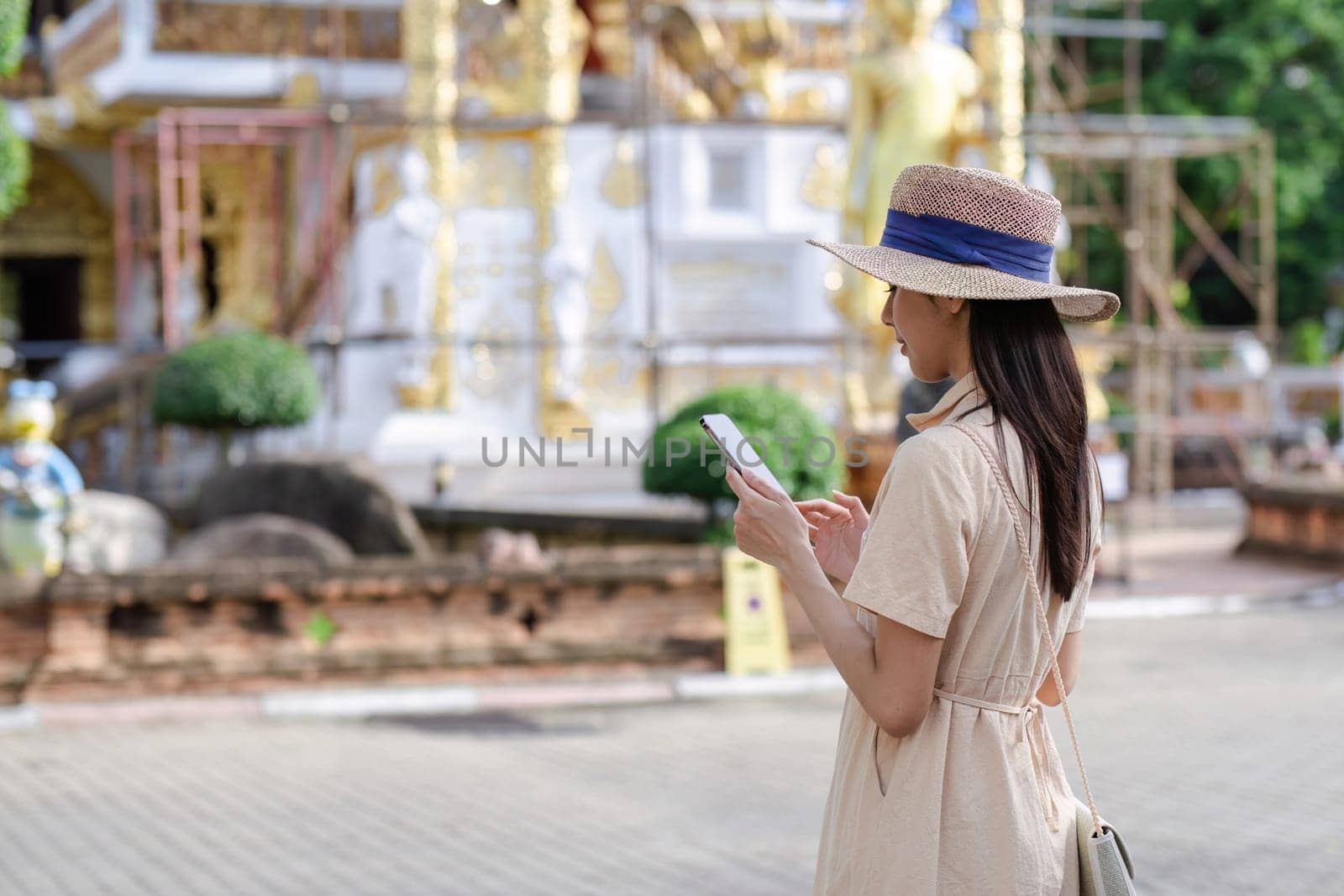 A young female tourist in a hat and dress uses her mobile phone while exploring a historic tourist attraction.