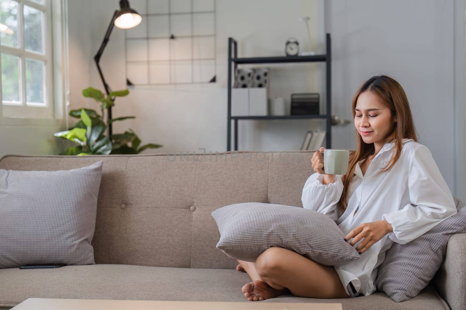 A young woman enjoys a peaceful morning, sitting on a couch with a cup of coffee in a modern, sunlit living room.