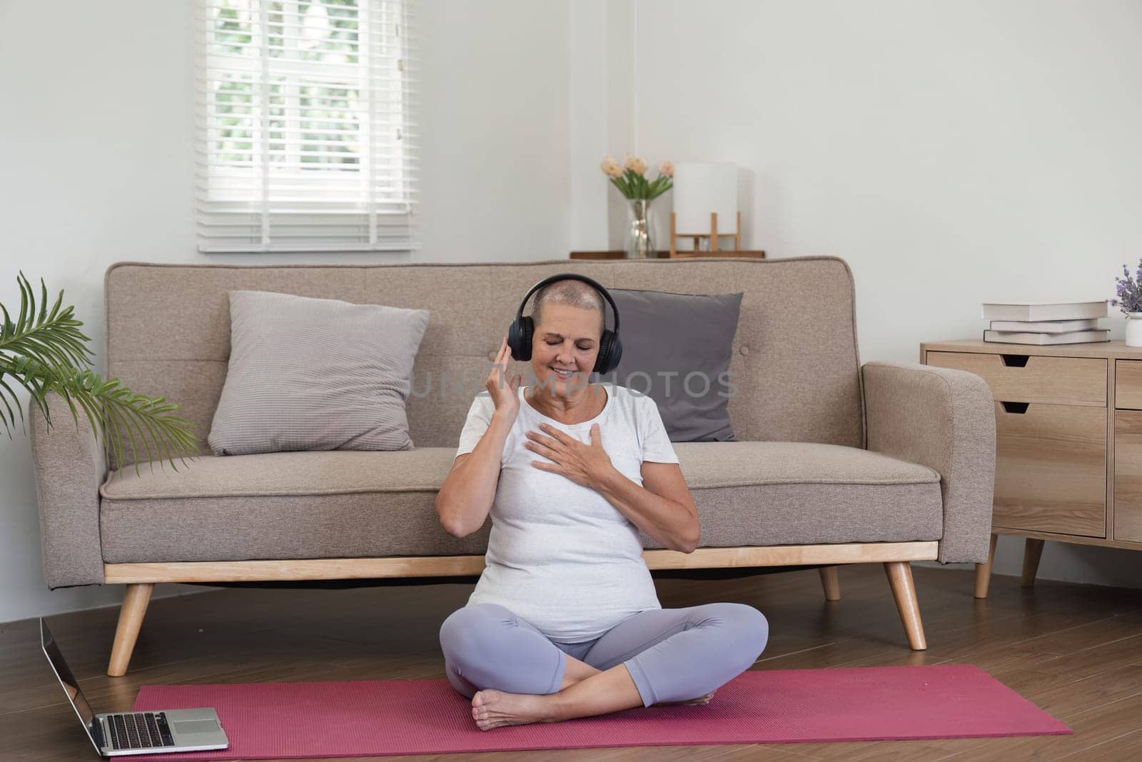 Elderly woman wearing headphones, sitting on a yoga mat, enjoying relaxing music in a modern living room setting.