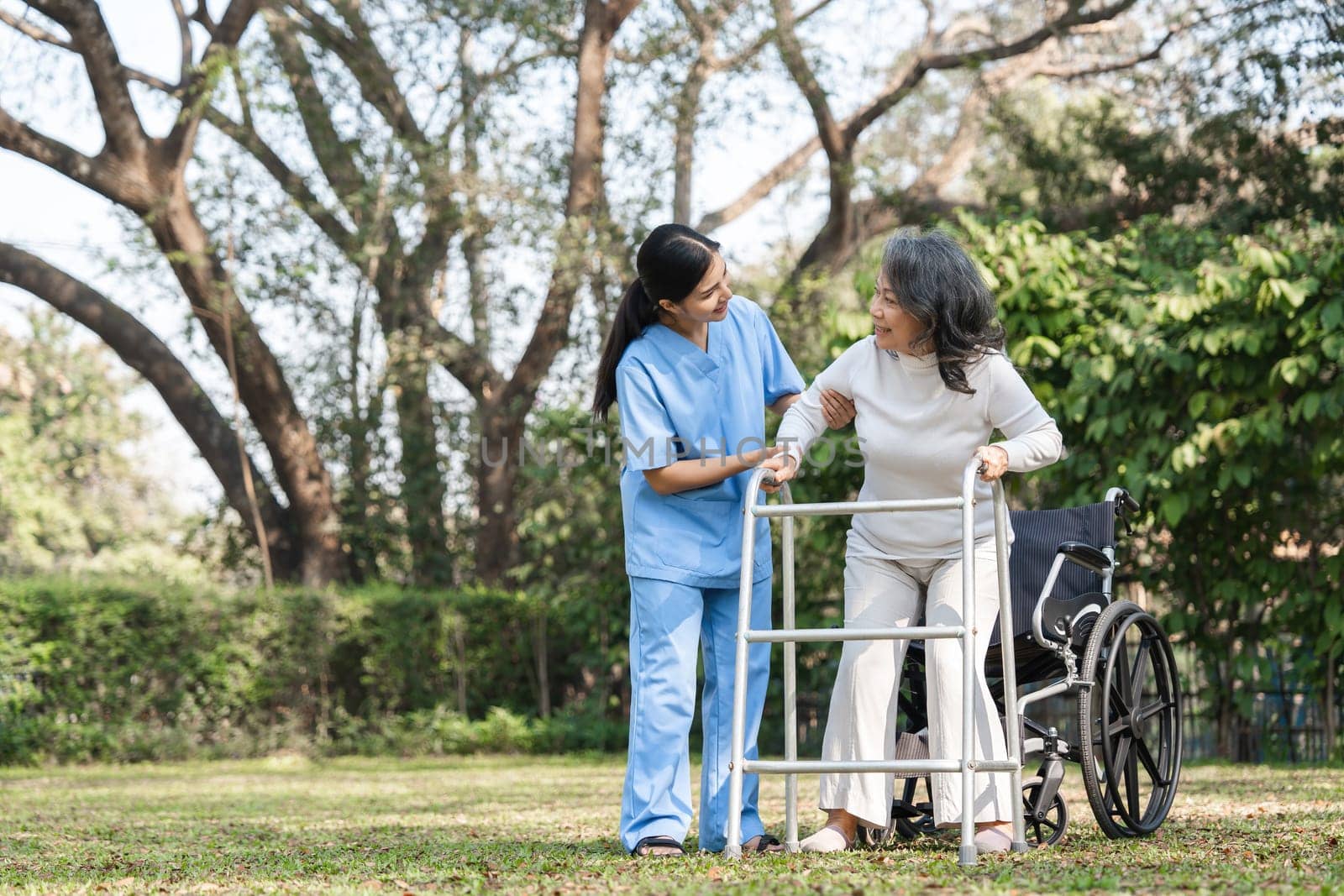 Caregiver helping an elderly woman with her rehabilitation exercises outdoors using a walker, promoting health and recovery.