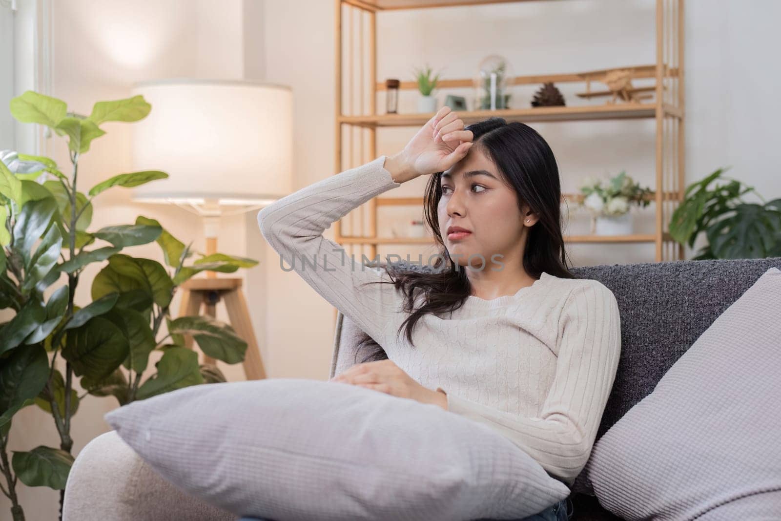 A young woman sitting on a sofa at home, holding her forehead and looking concerned, indicating she is feeling unwell or sick.
