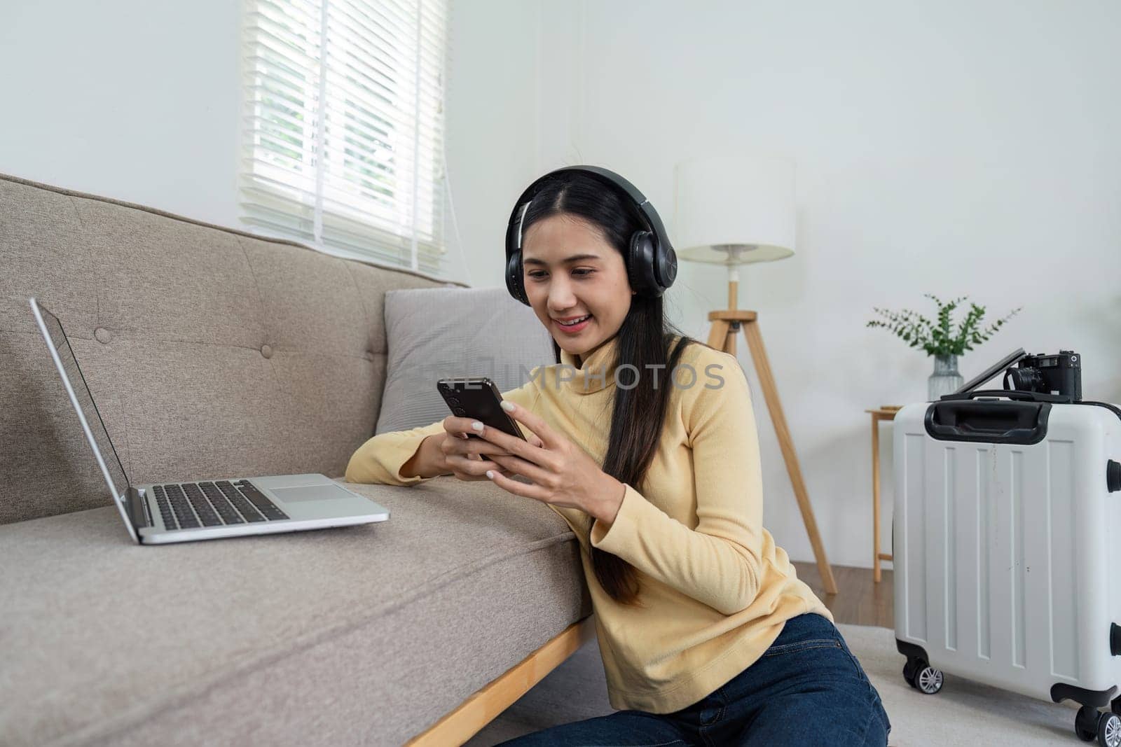 A young girl wearing headphones listens to music while using her smartphone and laptop in a comfortable living room setting.
