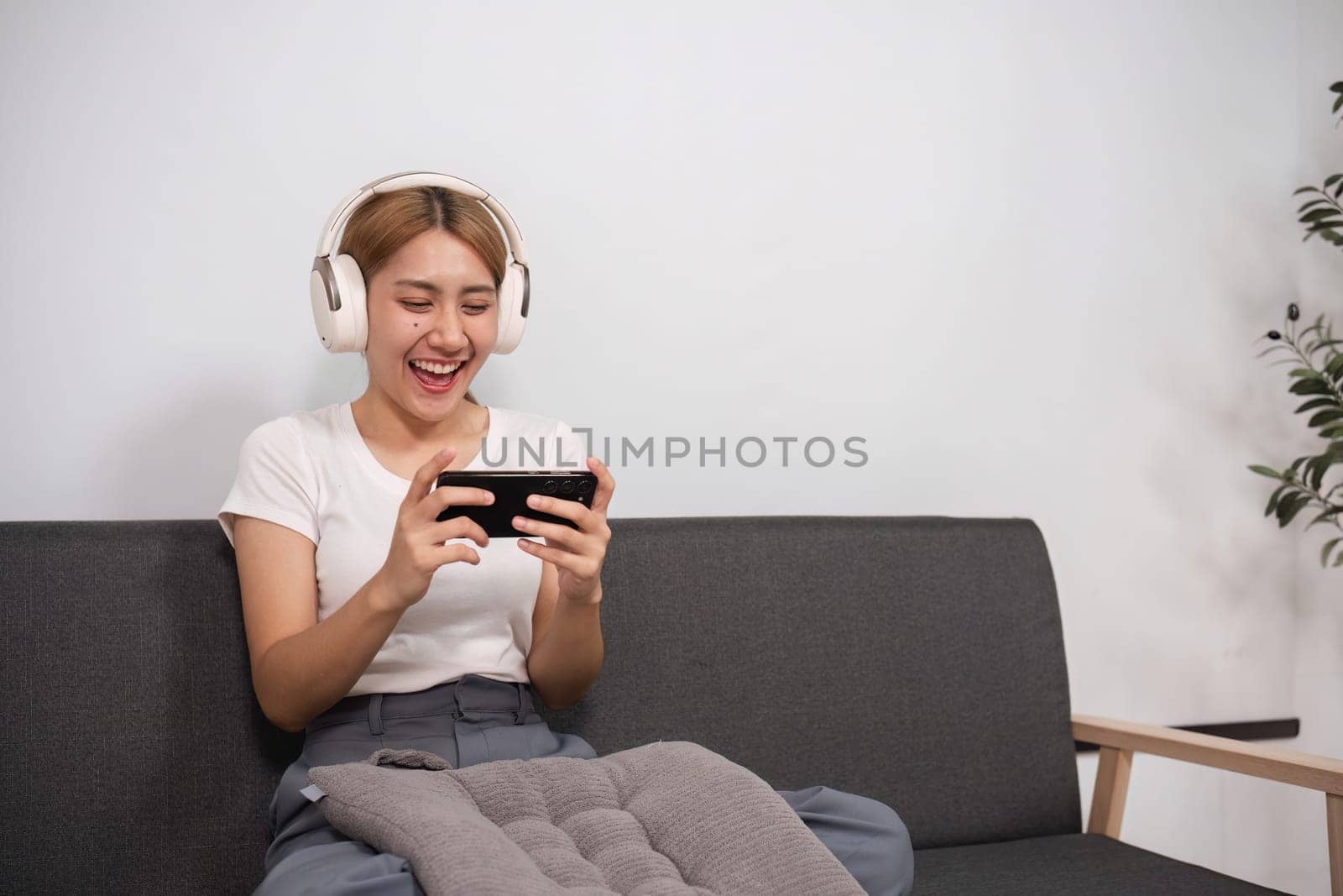 A young woman sits comfortably in a modern living room, wearing headphones and using a smartphone, enjoying a moment of relaxation.