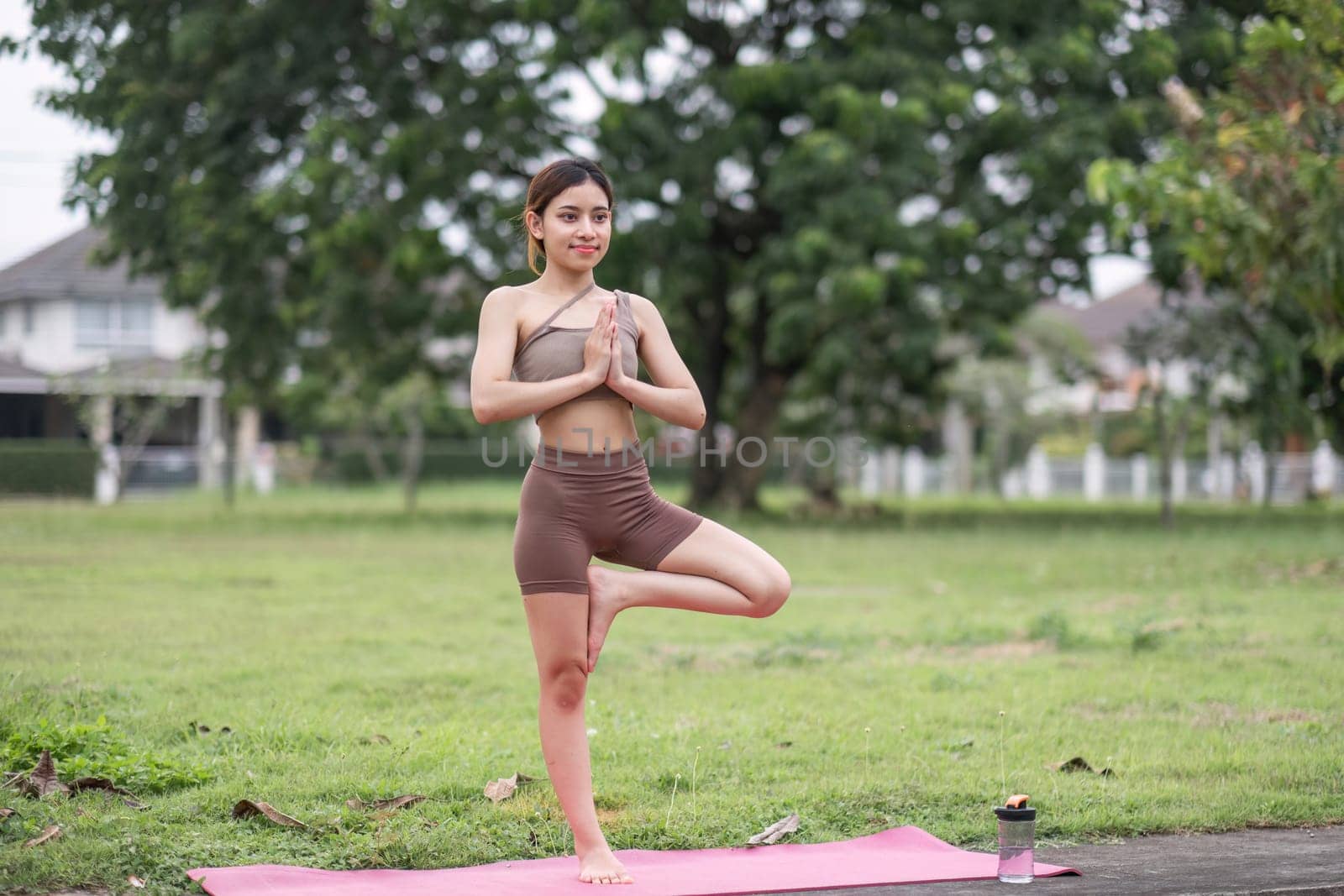A young woman performs yoga in a park, standing in tree pose on a pink mat, surrounded by lush greenery and trees.