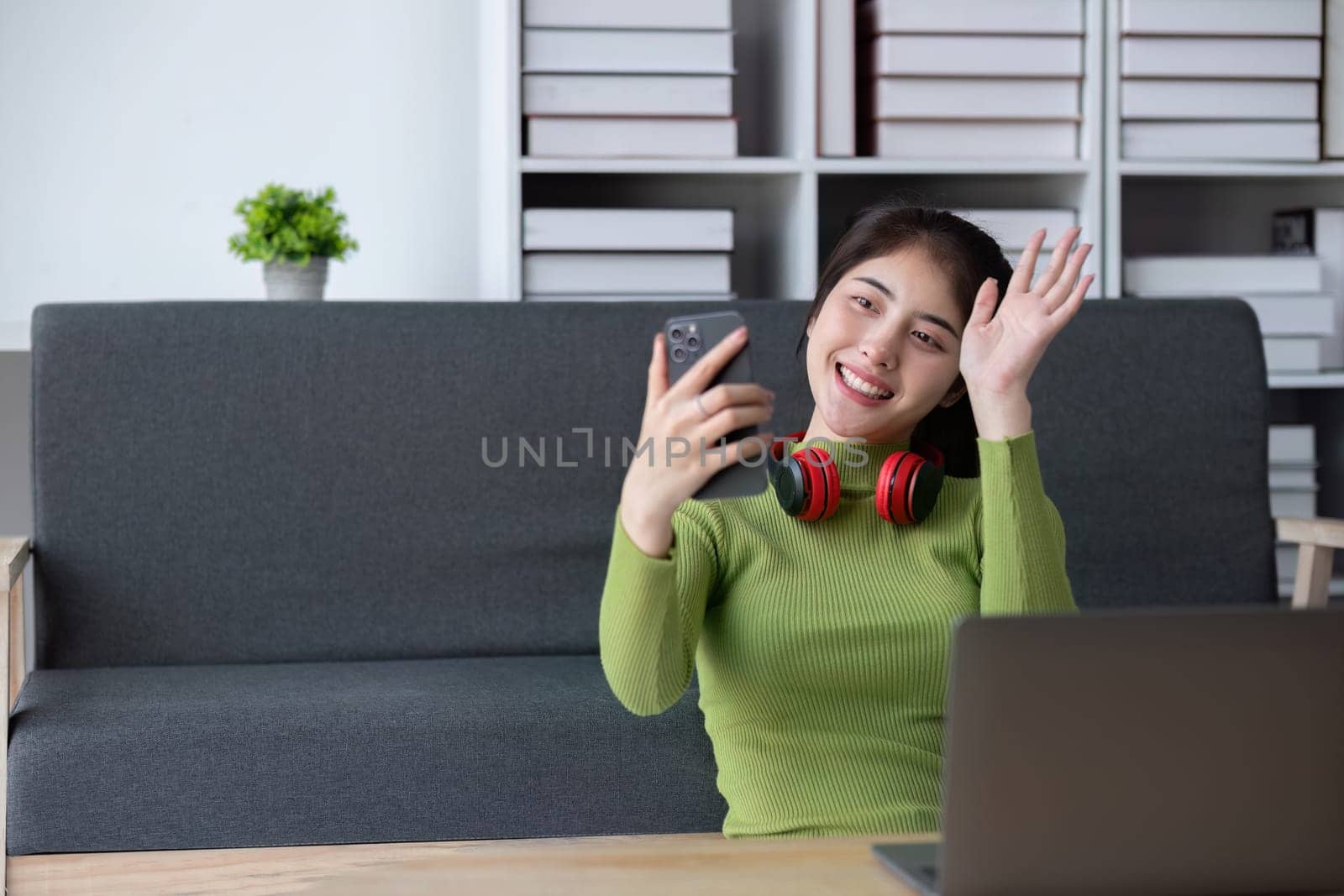 A cheerful young woman in a green sweater relaxes in a modern living room, using her smartphone and wearing red headphones.
