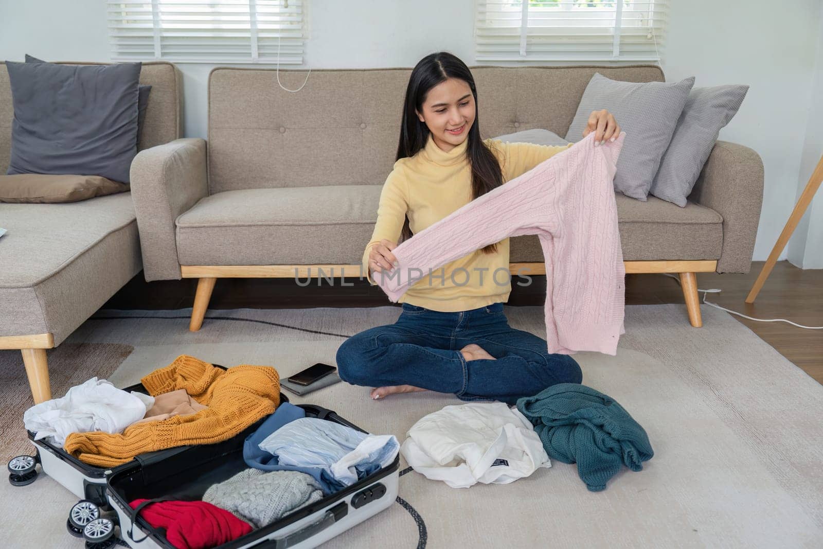 A young woman sits on the floor in a modern living room, packing a suitcase with clothes and essentials, preparing for travel or vacation.