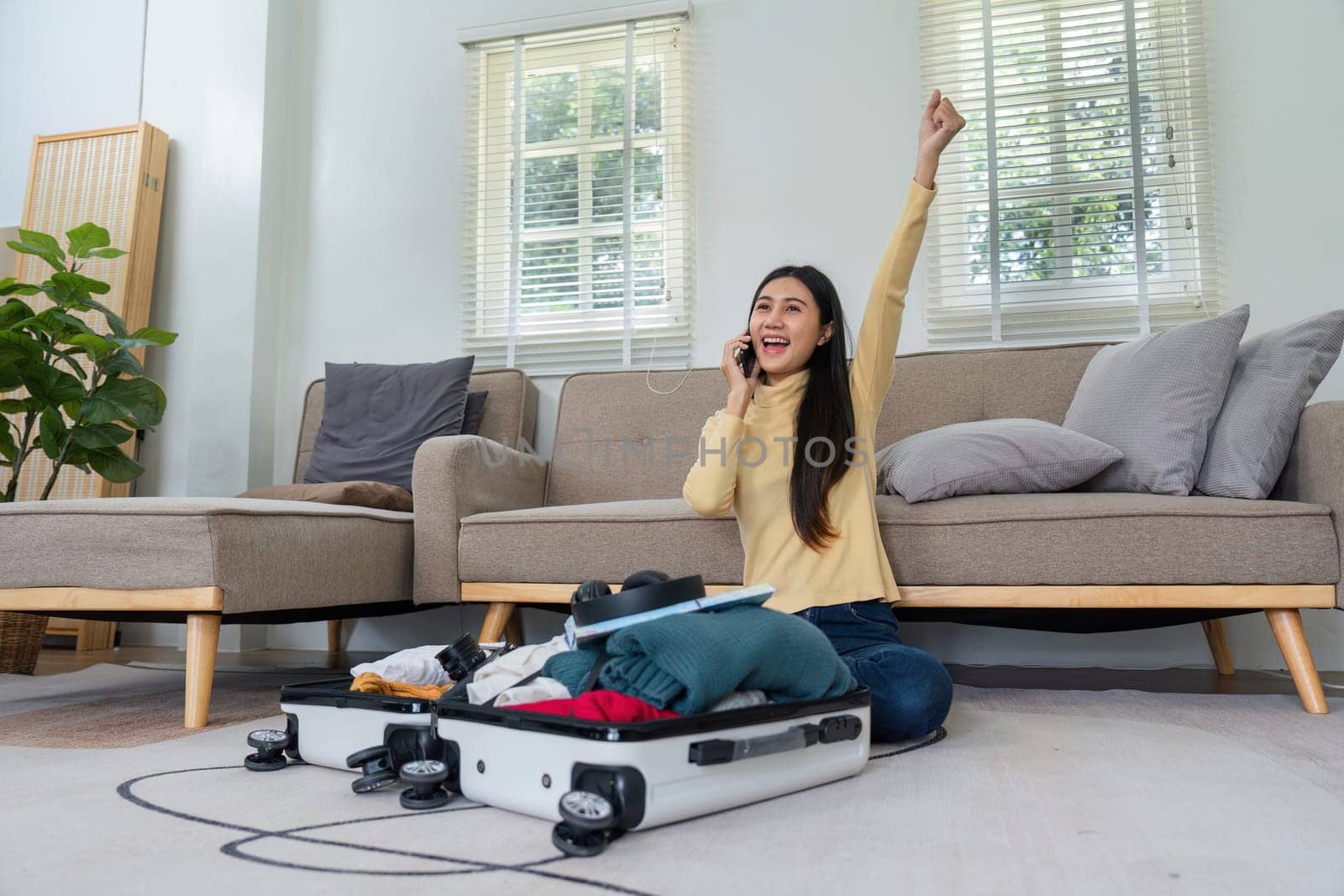 A cheerful young woman prepares her suitcase for travel while talking on the phone in a bright, modern living room.