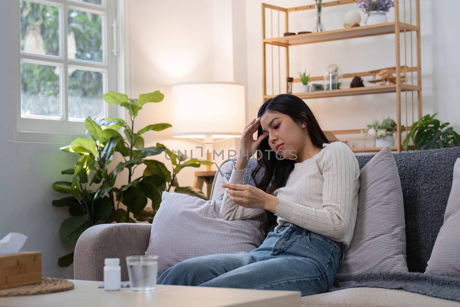 A young woman sitting on a couch, taking medicine while feeling unwell, in a cozy and modern living room with plants and soft lighting.