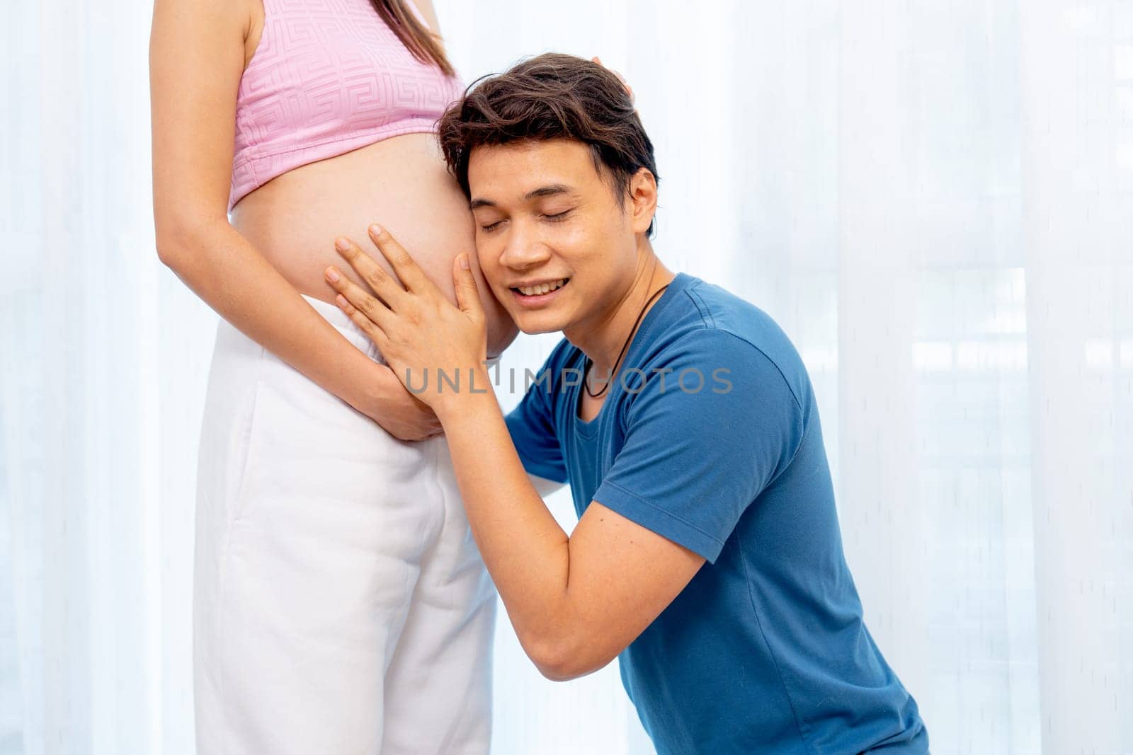 Asian man sit down and attach ear to belly of pregnant woman with smiling to express love and care in front of white curtain in their house.