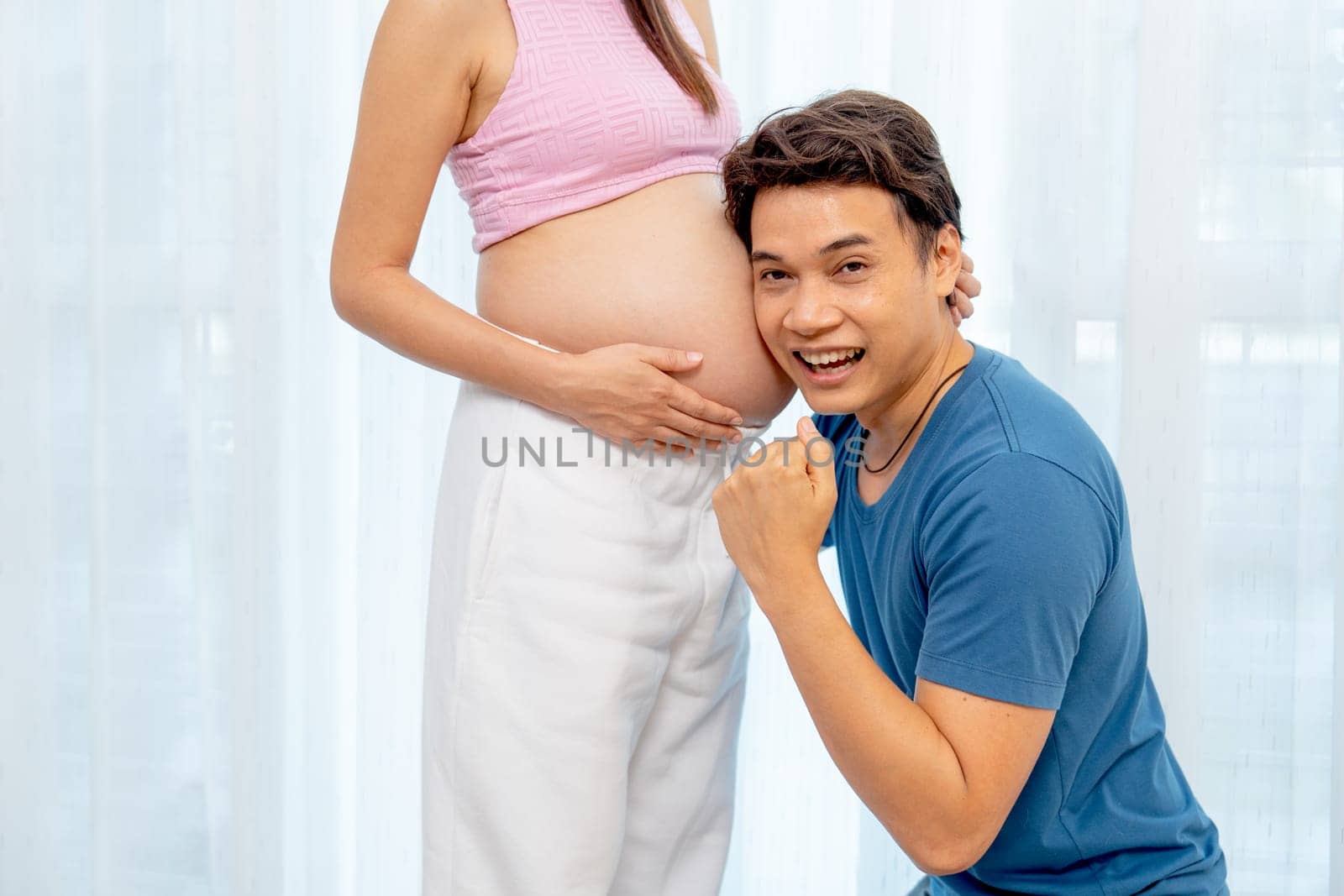 Asian man sit down and attach ear to belly of pregnant woman also look at camera show success emotion in front of white curtain in their house.
