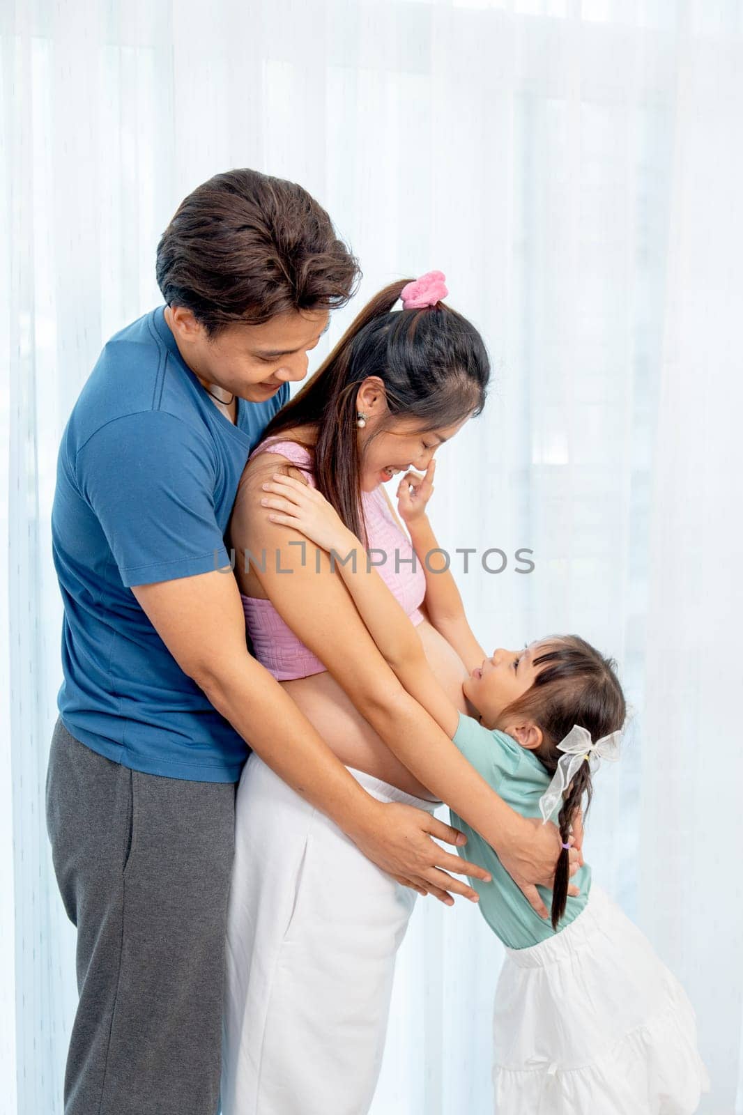 Asian family with father, pregnant mother and little girl hug together and stand in front of white curtain and girl touch mother nose.