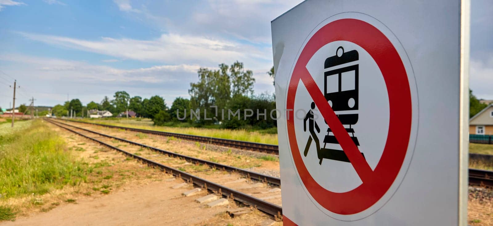 A concrete railway track under a sunny sky with a vivid prohibition sign displaying a train and a person, emphasizing no crossing. The scene captures rail safety on a lovely day.