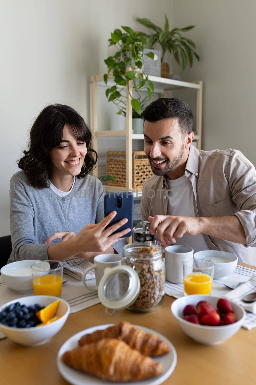 Woman showing man phone while eating breakfast at home. Happy couple having healthy meal at home looking at mobile phone. Vertical image. Lifestyle concept.