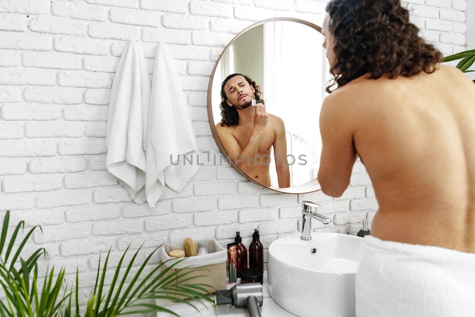 A man with curly hair grooming himself in the bathroom while looking into a round mirror close up