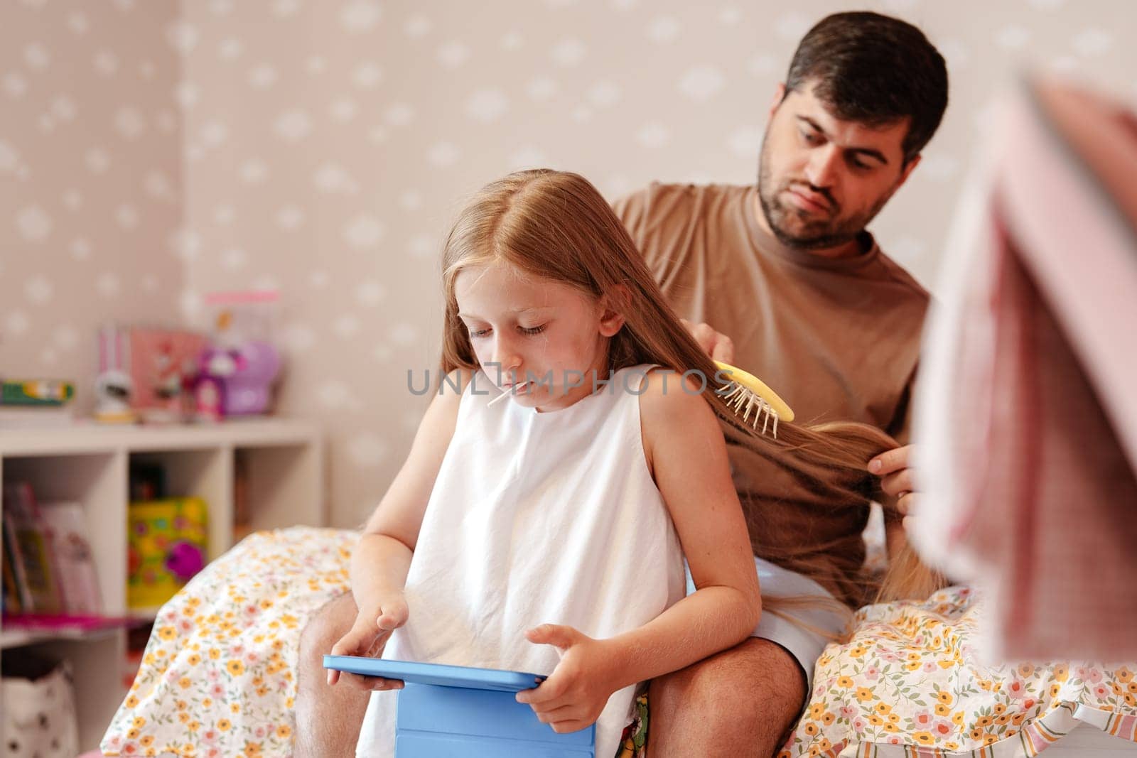 A man sits on a bed and brushes a young girl's hair. The girl looks at a tablet in her hands. They are in a bedroom with a white dresser in the background.