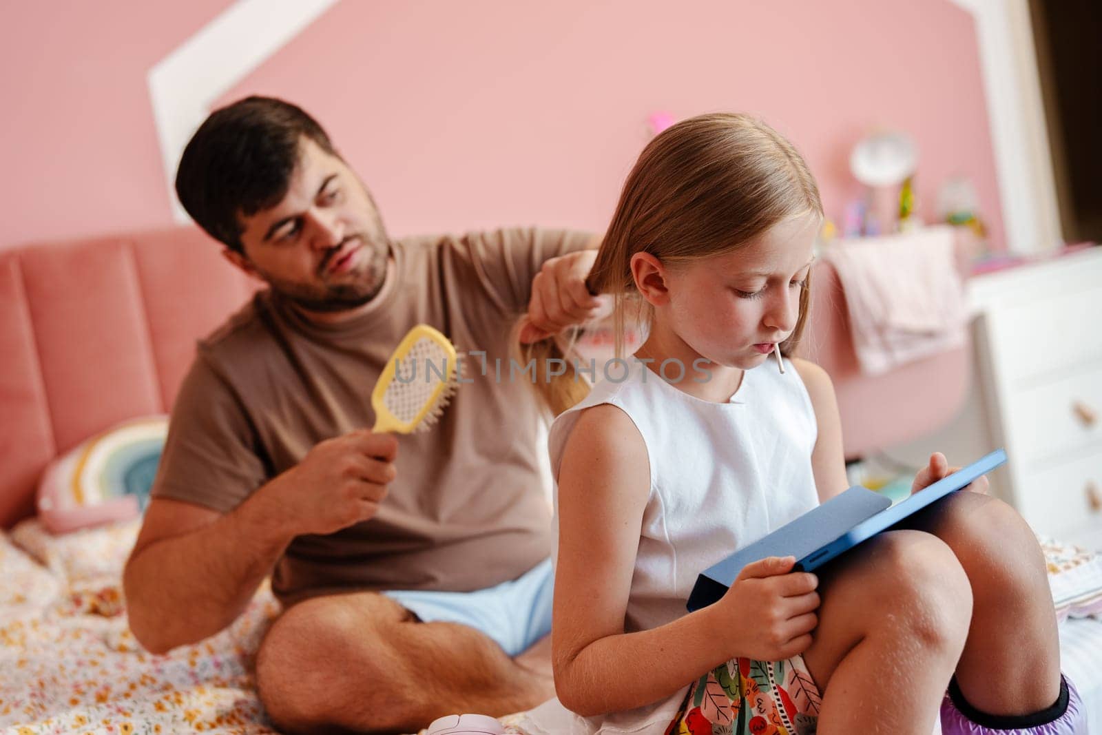 A man sits on a bed and brushes his daughter's hair. She is sitting beside him, reading a book. She is focused on the book while he brushes her hair.