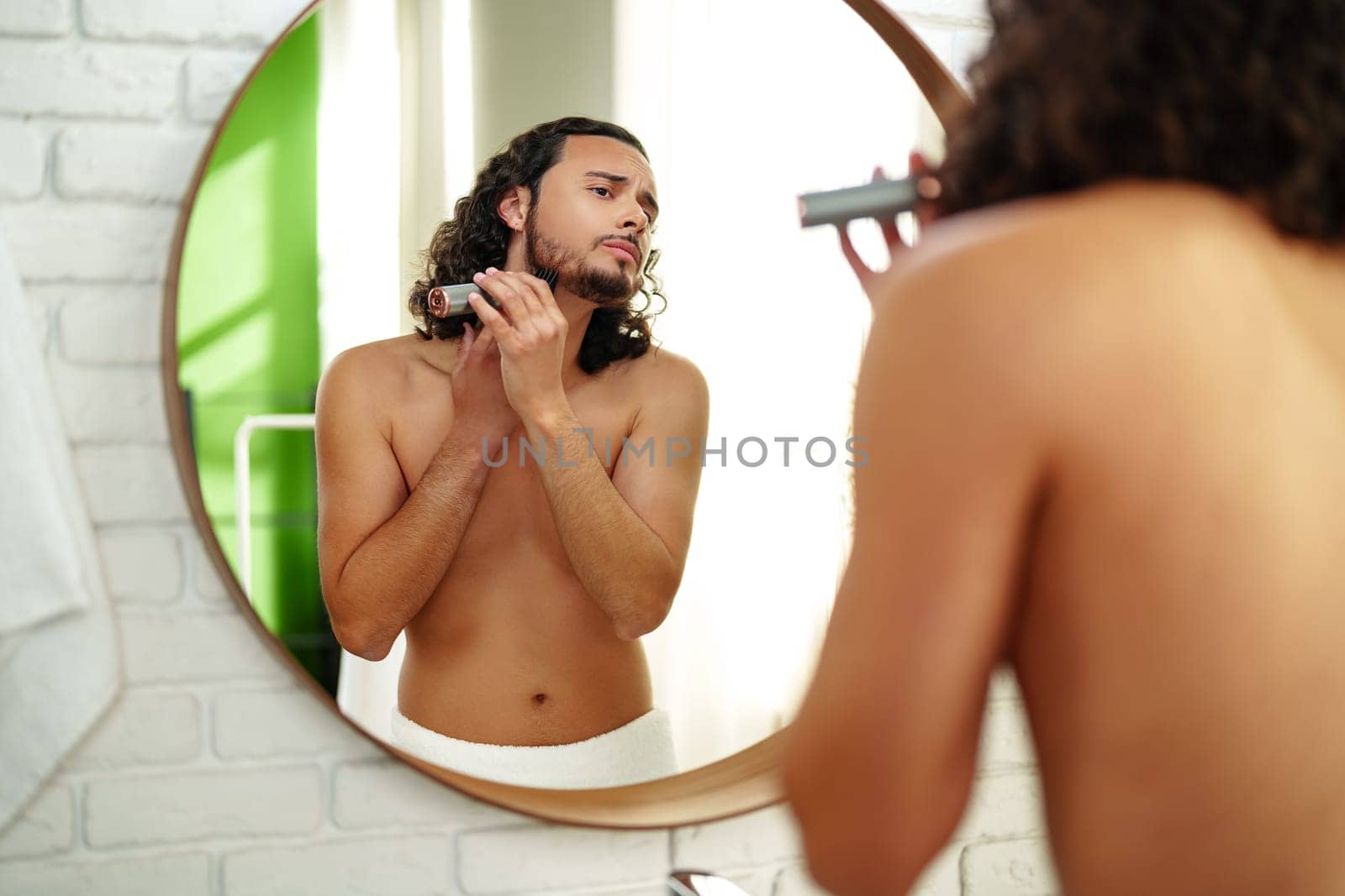 A man with curly hair grooming himself in the bathroom while looking into a round mirror close up