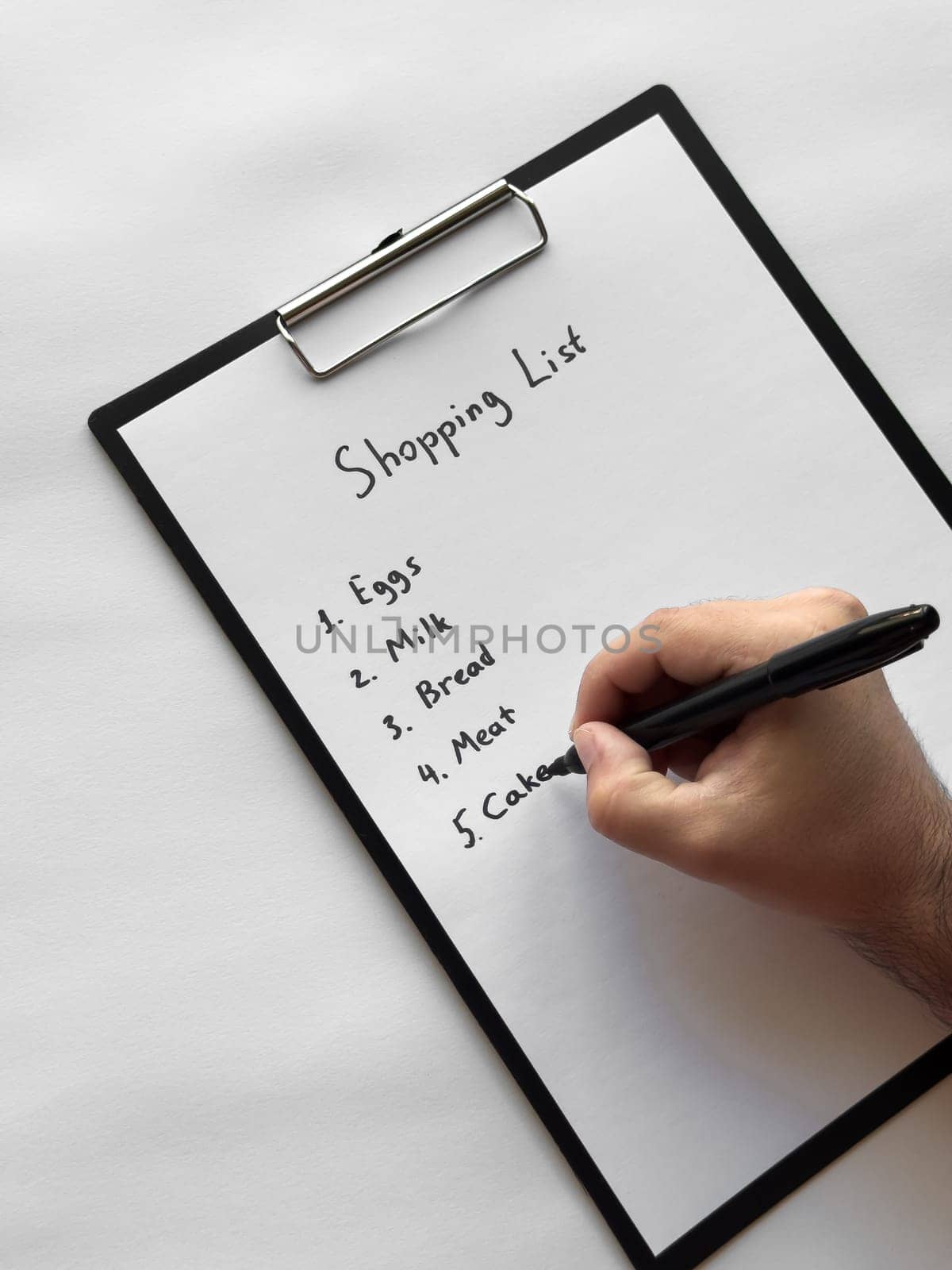 Hand writing shopping list on clipboard with eggs, milk, bread, meat, cakes items.