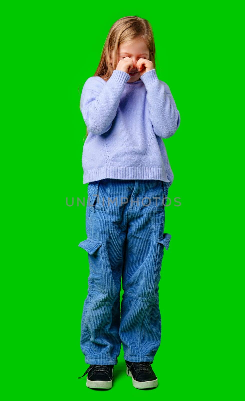 A young girl in a cozy sweater and blue cargo pants expressing emotion with a bright green background in studio