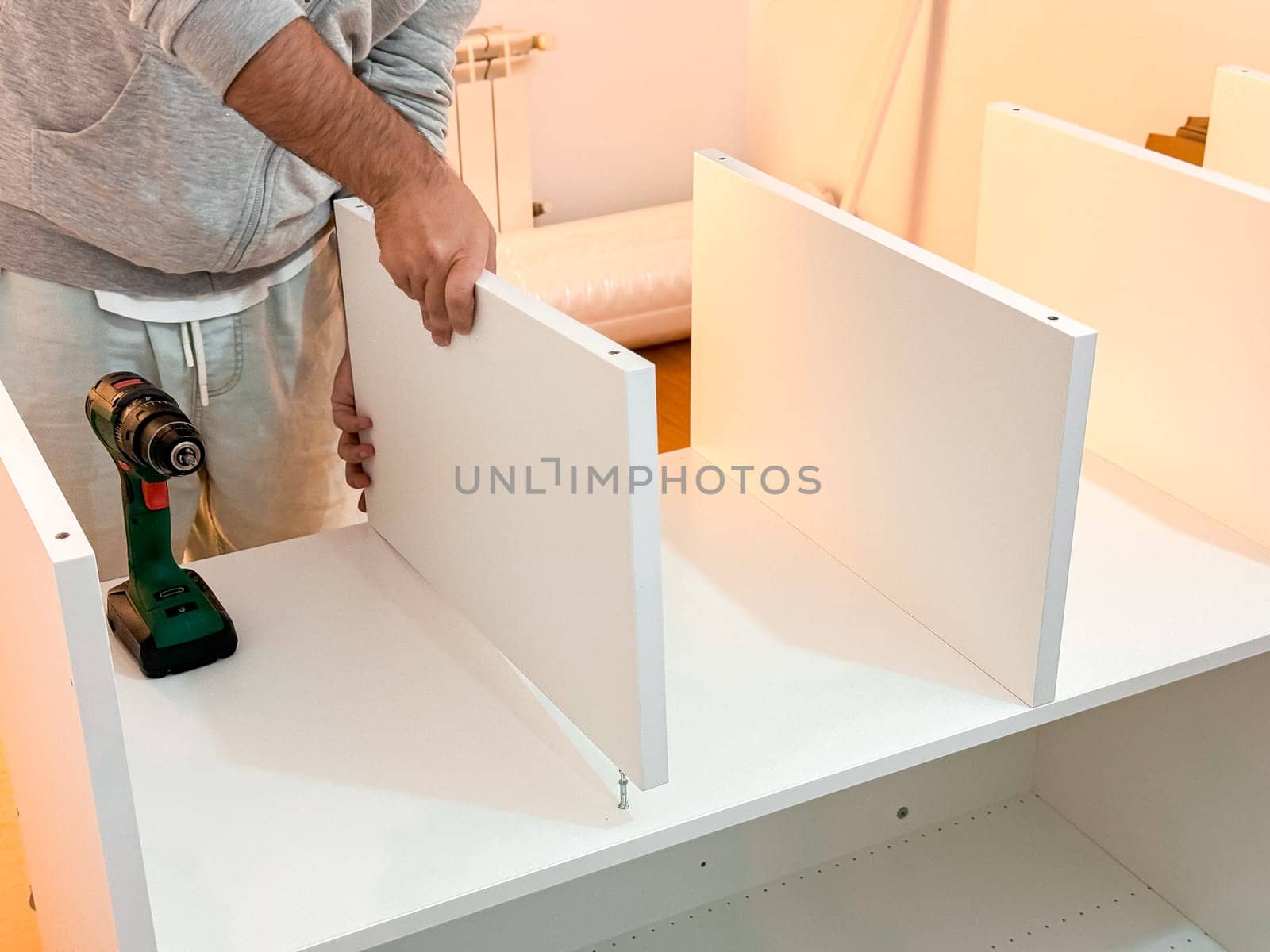 Caucasian male adult assembling white shelving unit with electric drill at home.