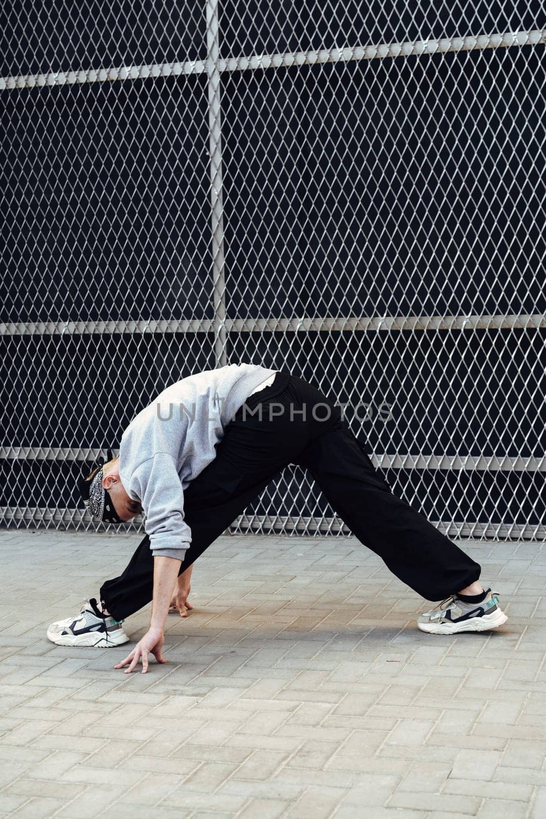 A person wearing a gray sweatshirt and black pants is stretching on pavement in front of a chain link fence. They are leaning forward, reaching their hands down to touch the ground.