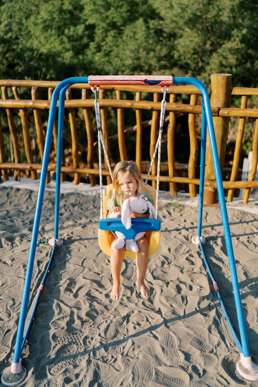 Little smiling girl swinging on a swing hugging a pink bunny at the playground. High quality photo