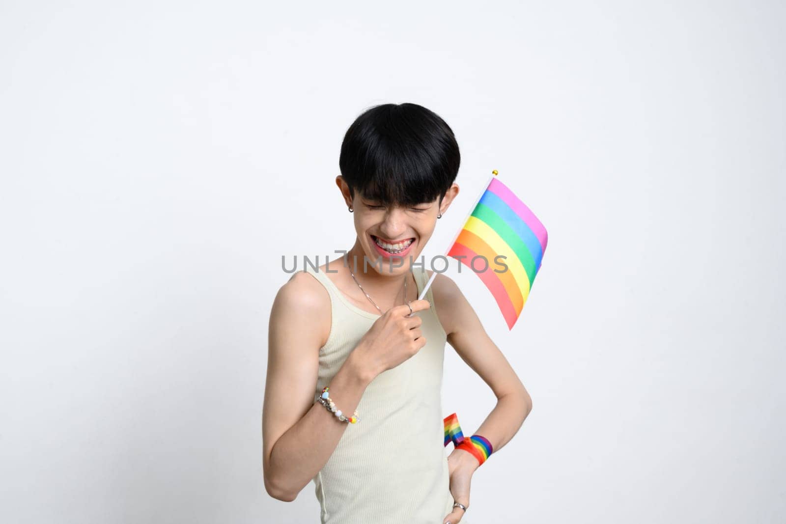Smiling young man holding a small rainbow pride flag over white background to celebrating LGBTQ pride.