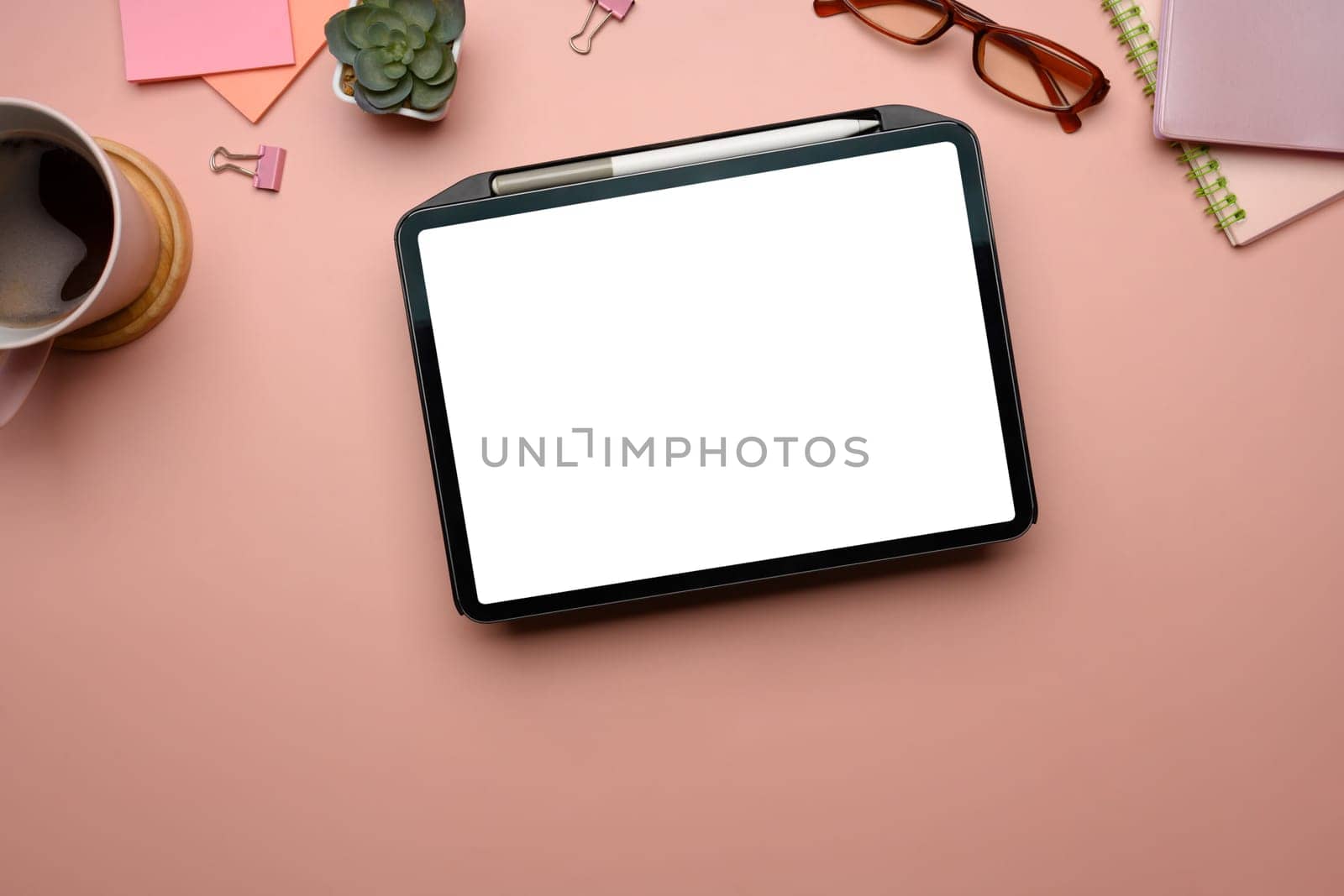 Feminine workspace with digital tablet, coffee cup, notebooks and accessories on pastel pink background.