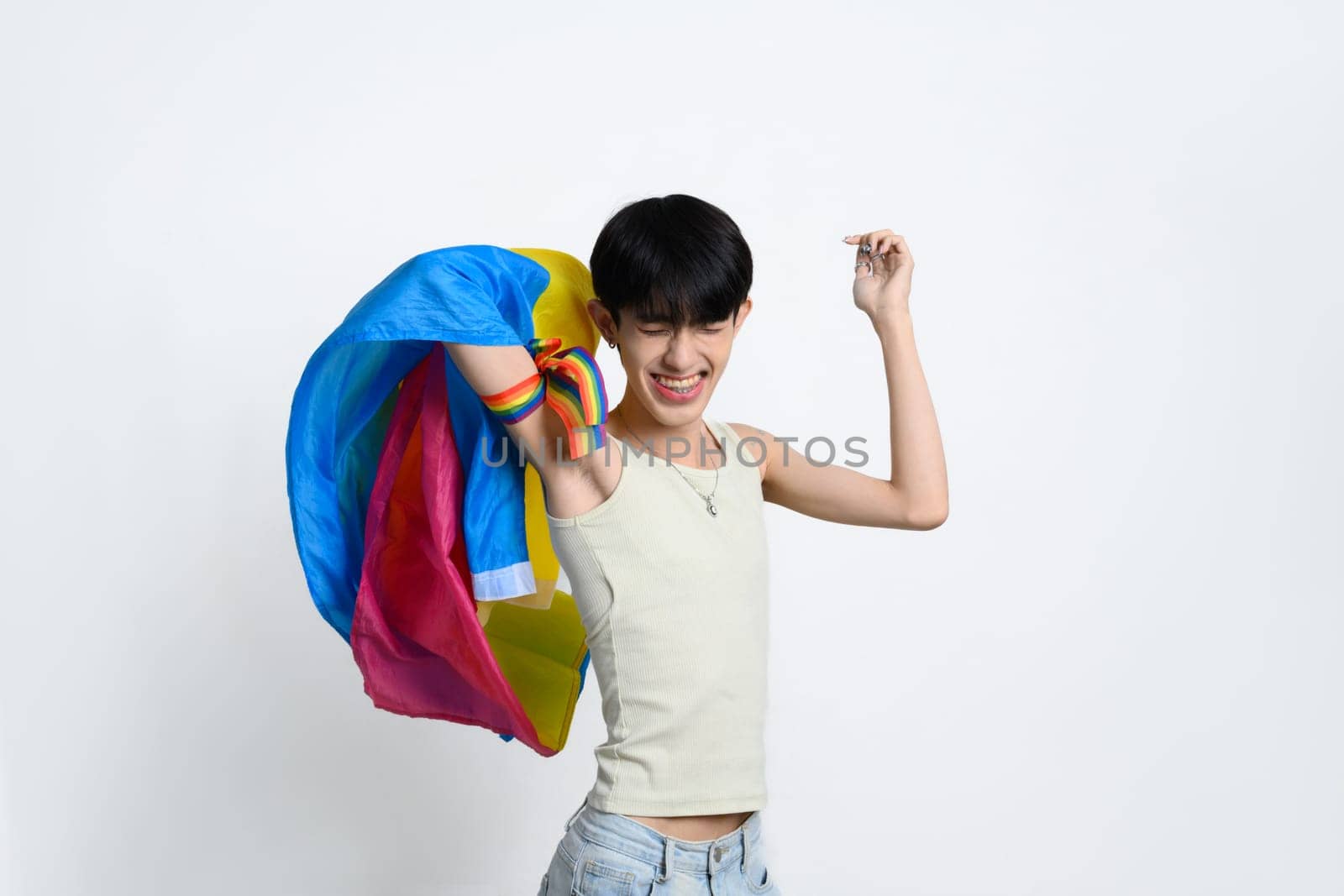 Cheerful teenage man joyfully posing with rainbow pride flag on white background. LGBTQ pride and equality concept.