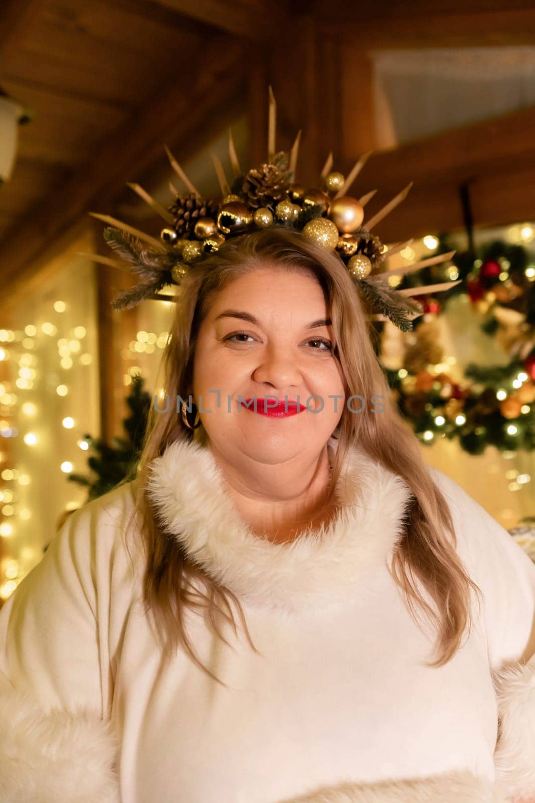 A woman wearing a white sweater and a gold crown with ornaments smiles at the camera. A Christmas wreath is visible in the background.
