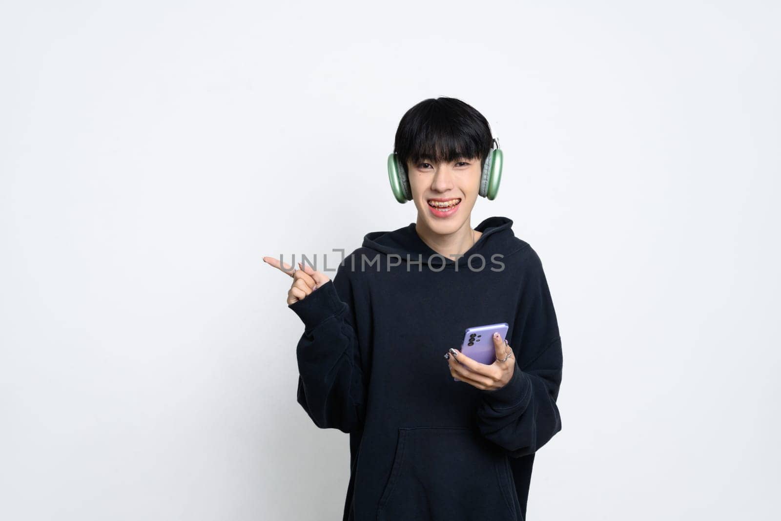 Cute asian man in headphone holding smartphone and pointing pointing aside to present a product.