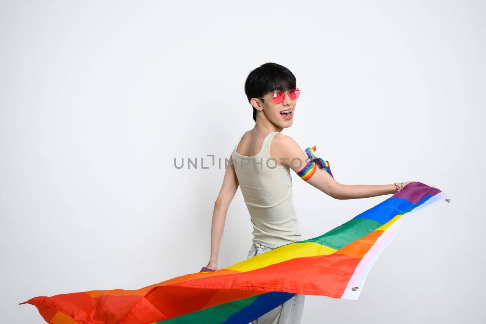 Joyful asian man wearing pink sunglasses swirling with pride flag against white background. LGBTQ pride and support.