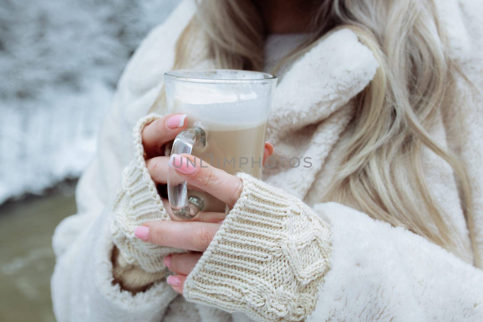 Beautiful hands hold a cup of cappuccino against the background of the winter forest. Gently beige gloves on the hands of a girl in the hands of coffee.